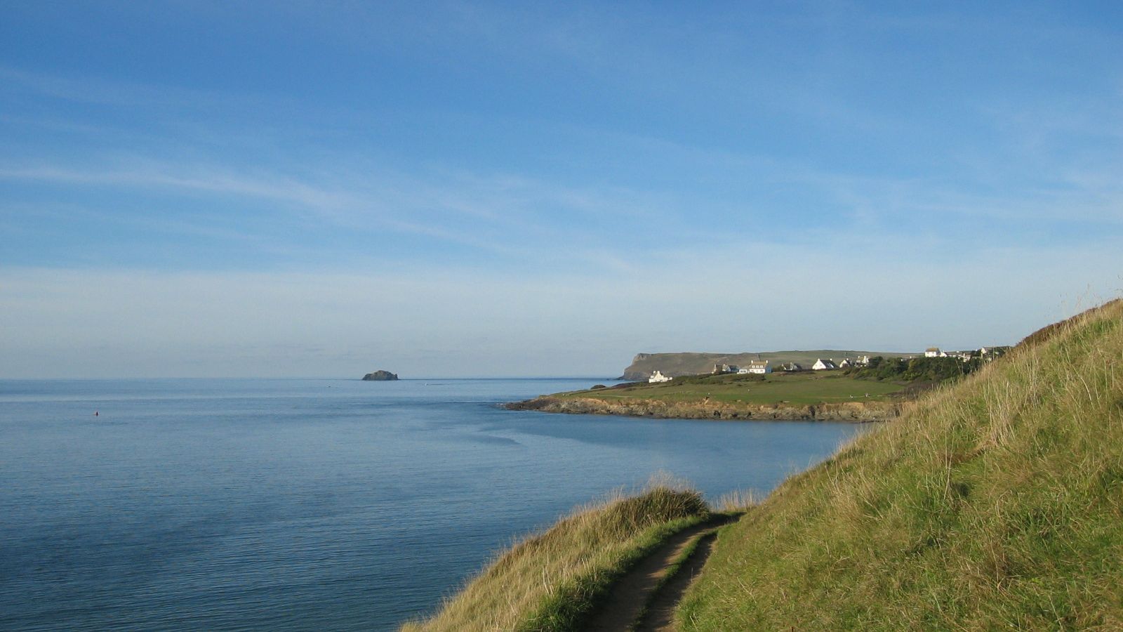 Coastal path along grassy hill overlooking calm sea with distant shoreline and houses
