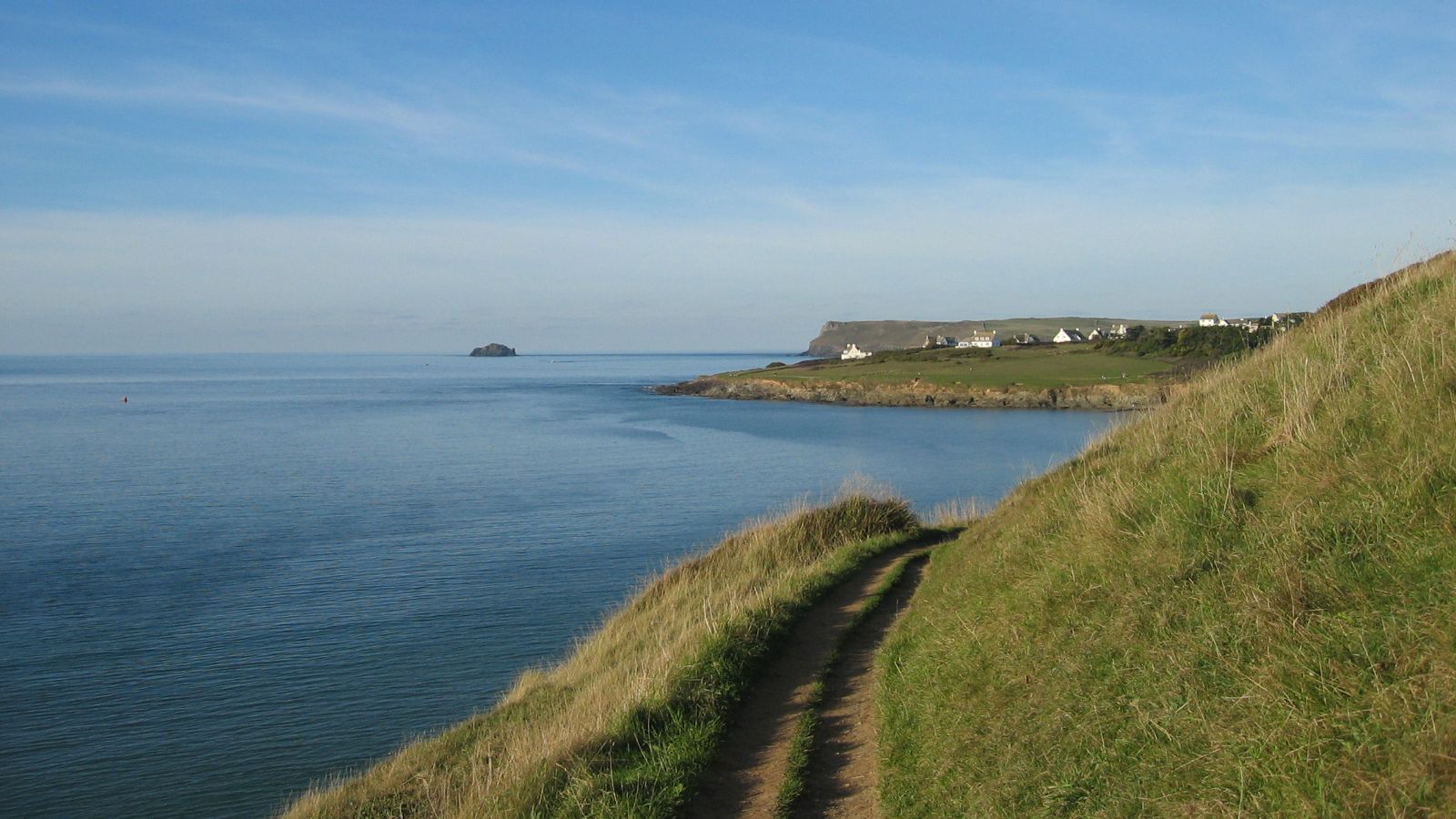 Coastal path along grassy hill overlooking calm sea with distant shoreline and houses