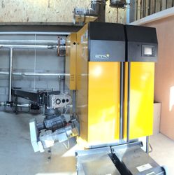 Interior of a biomass boiler room showing a red expansion vessel, a large white thermal storage tank, and a yellow biomass boiler unit with metal piping.