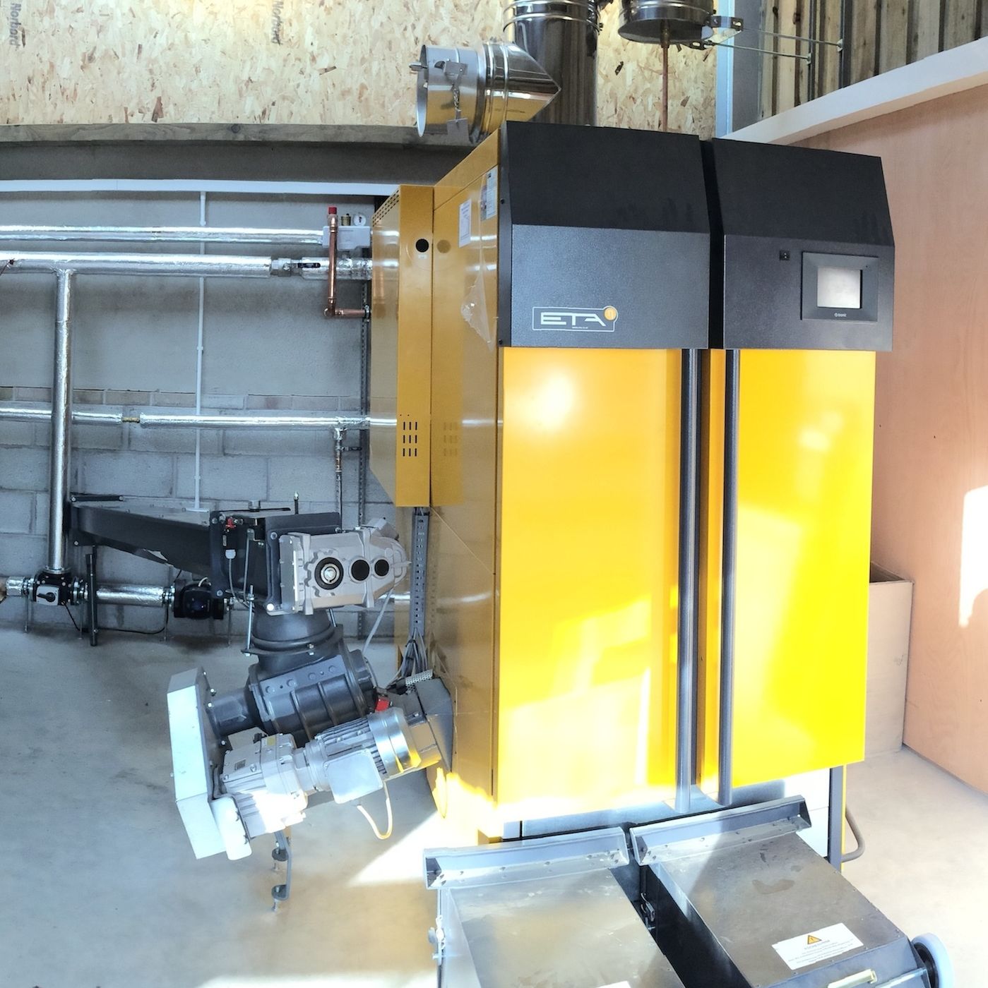 Interior of a biomass boiler room showing a red expansion vessel, a large white thermal storage tank, and a yellow biomass boiler unit with metal piping.