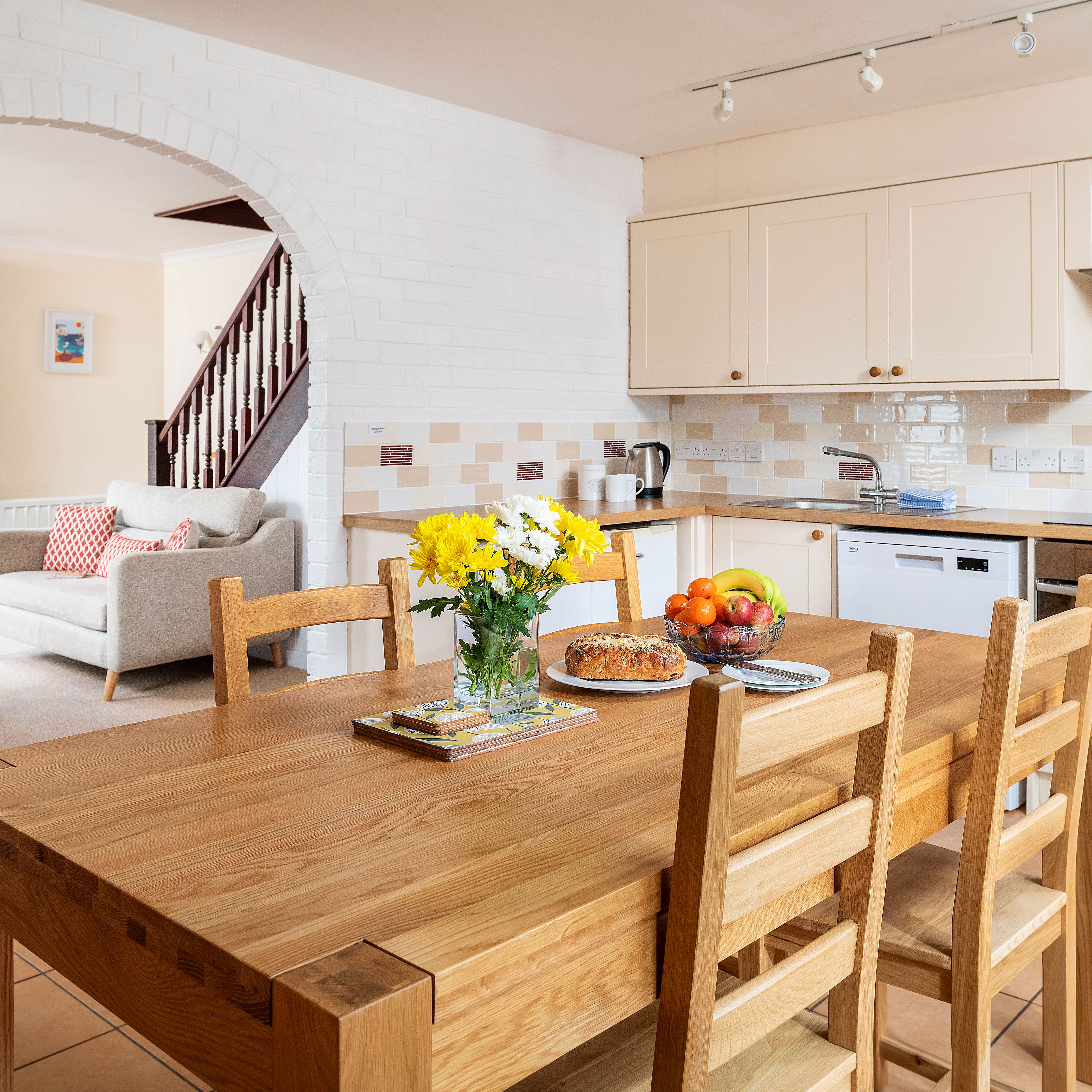 Modern kitchen and open living area with wooden dining table, fruit bowl, and flowers