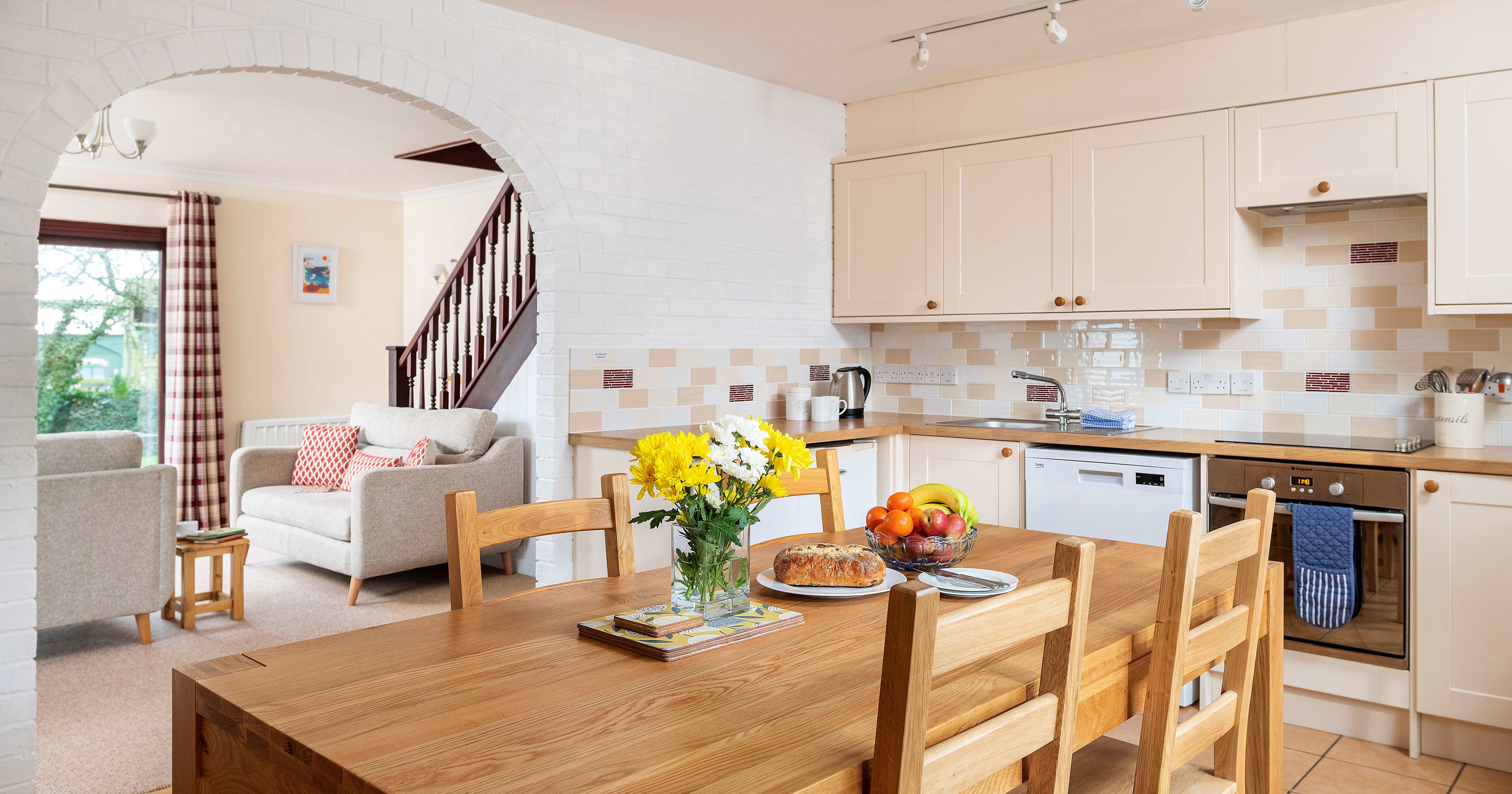 Modern kitchen and open living area with wooden dining table, fruit bowl, and flowers