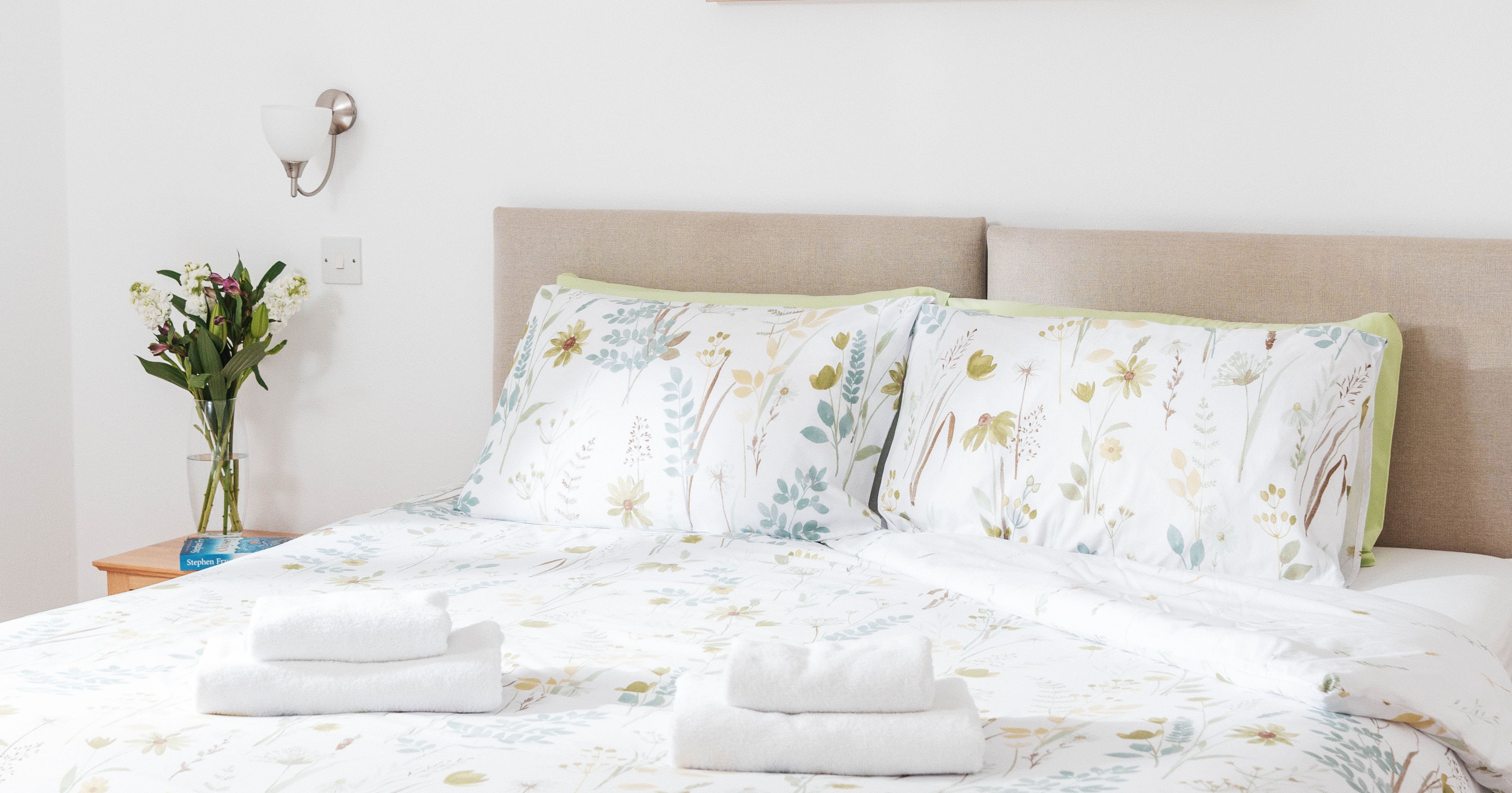 Bright bedroom with a double bed, floral bedding, two sets of folded towels, wall art, and a vase of flowers on a bedside table.