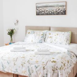 Bright bedroom with a double bed, floral bedding, two sets of folded towels, wall art, and a vase of flowers on a bedside table.