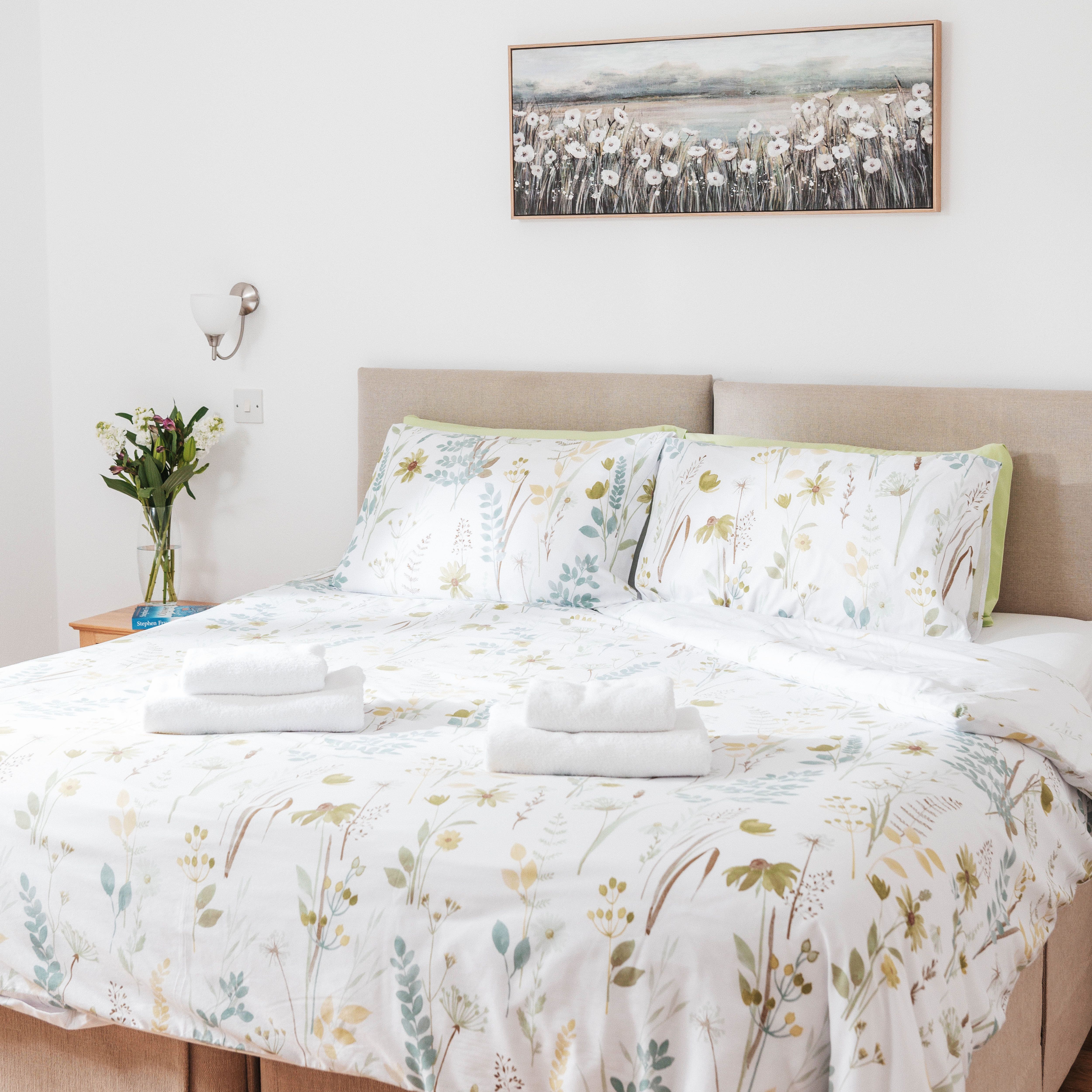 Bright bedroom with a double bed, floral bedding, two sets of folded towels, wall art, and a vase of flowers on a bedside table.