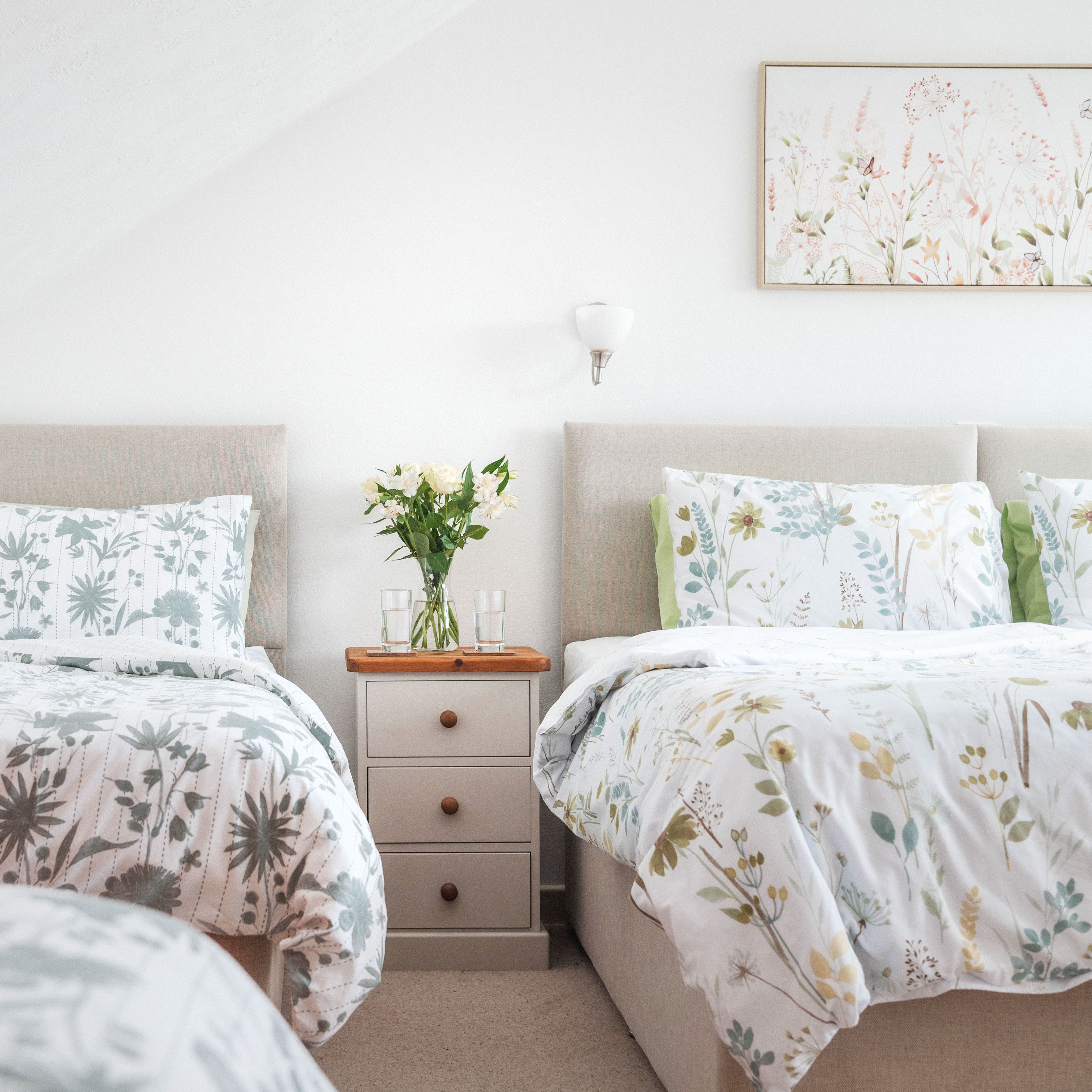 Cozy bedroom with two beds, floral bedding, a wooden dresser with books, and a nightstand with flowers and glasses of water