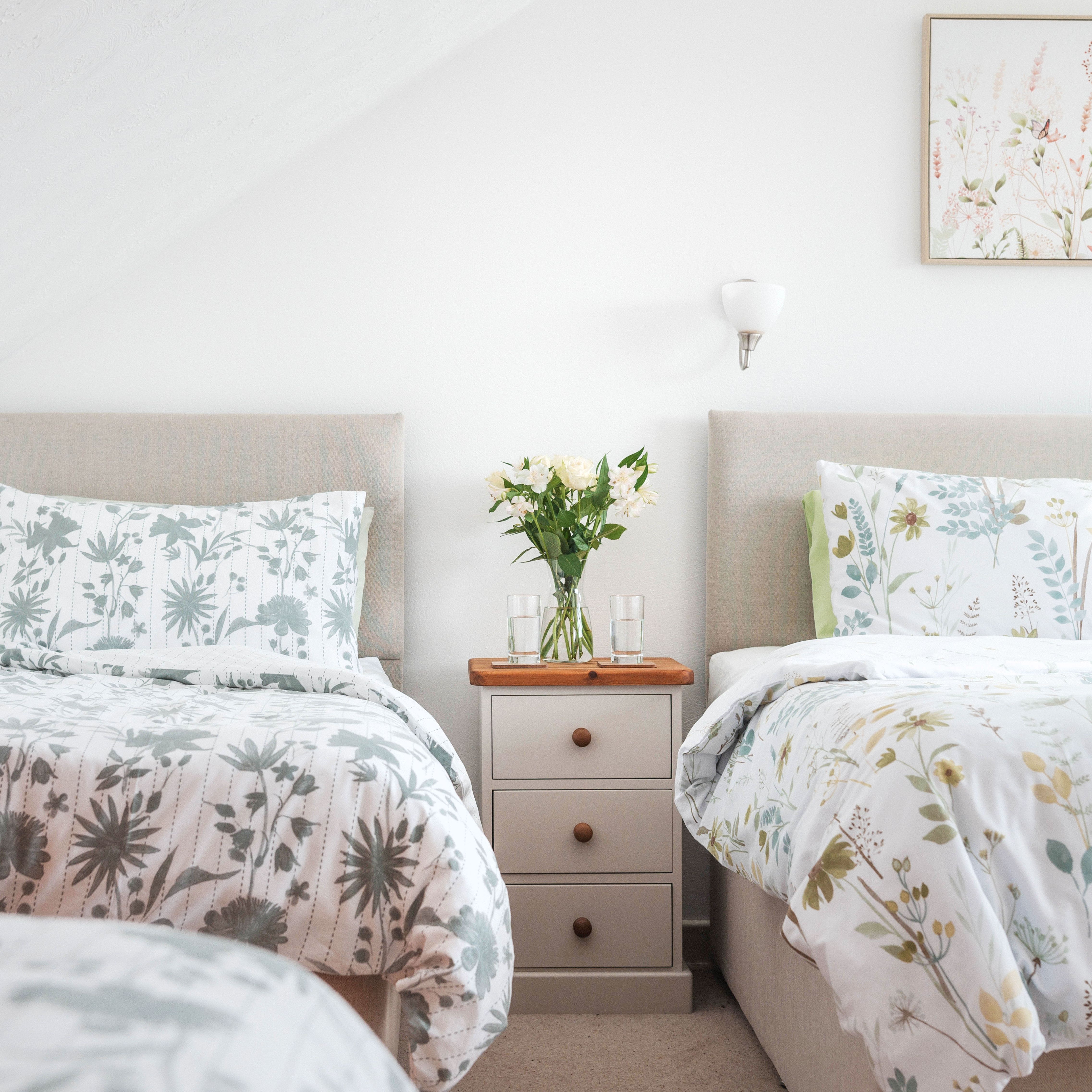 Cozy bedroom with two beds, floral bedding, a wooden dresser with books, and a nightstand with flowers and glasses of water