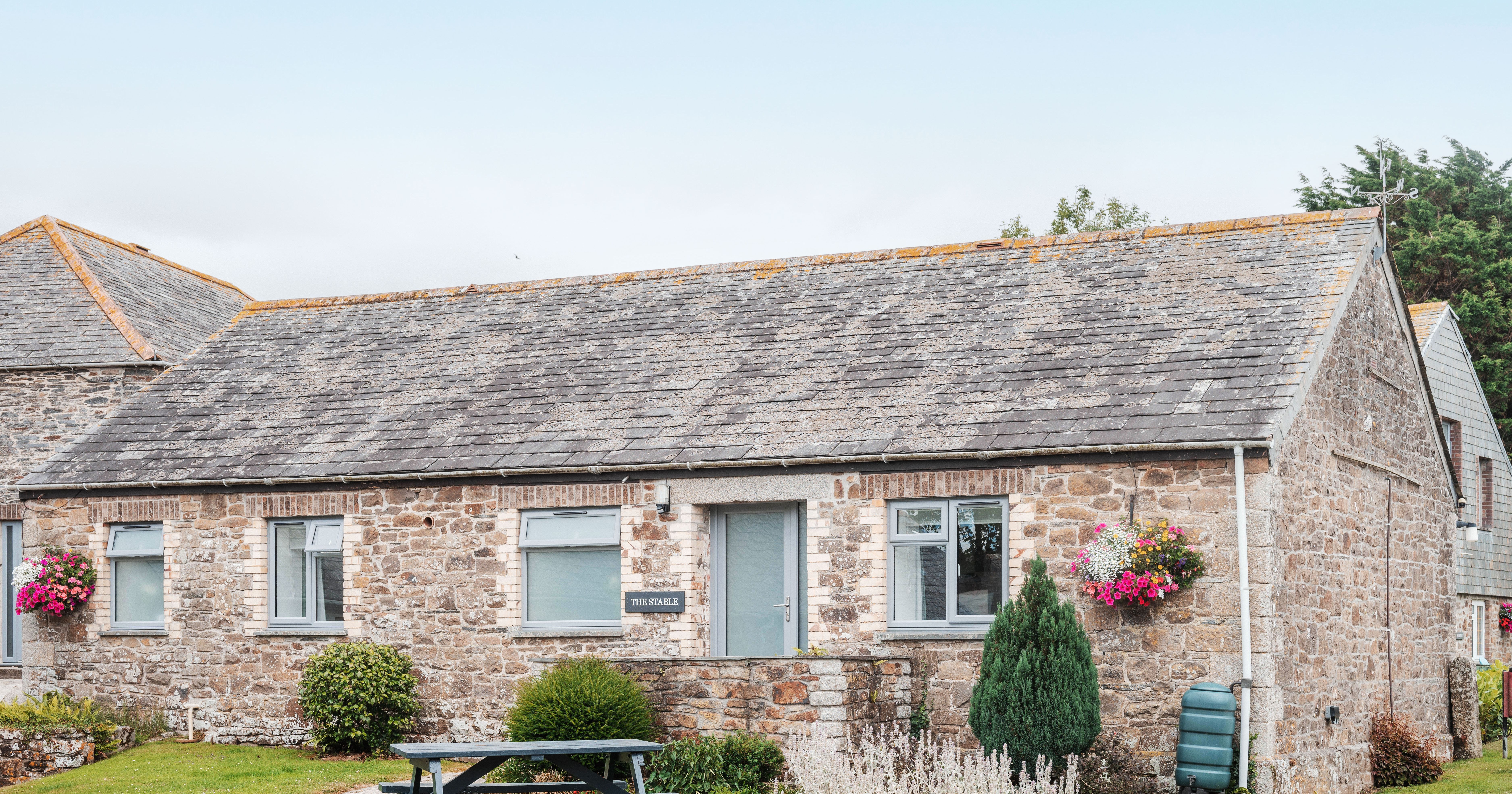 Stone cottage with slate roof, hanging flower baskets, green bushes, and a picnic table outside.