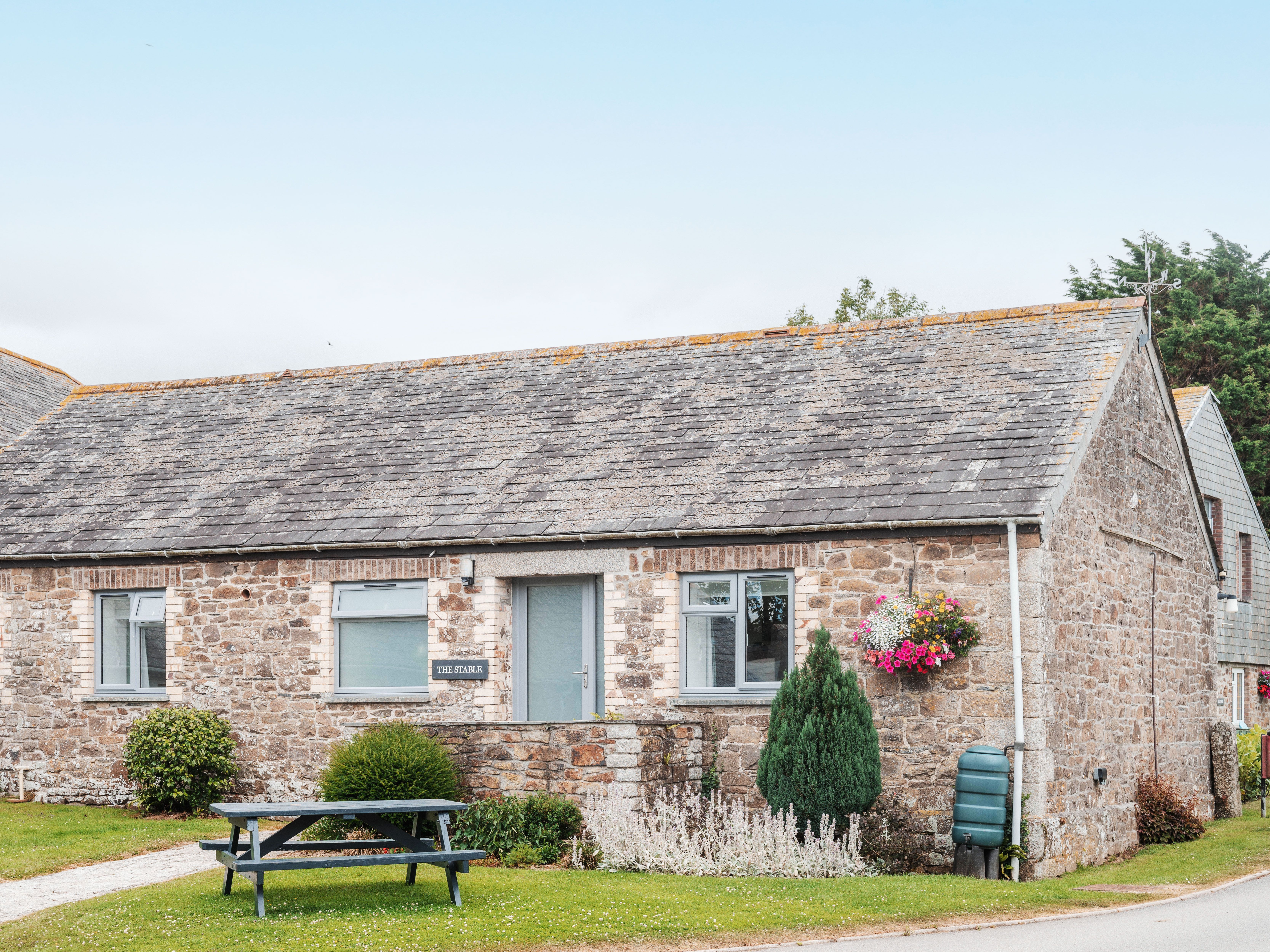 Stone cottage with slate roof, hanging flower baskets, green bushes, and a picnic table outside.