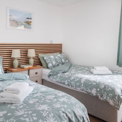 Two twin beds with floral bedding, bedside tables with lamps, and a window with green curtains in a bright, modern bedroom.