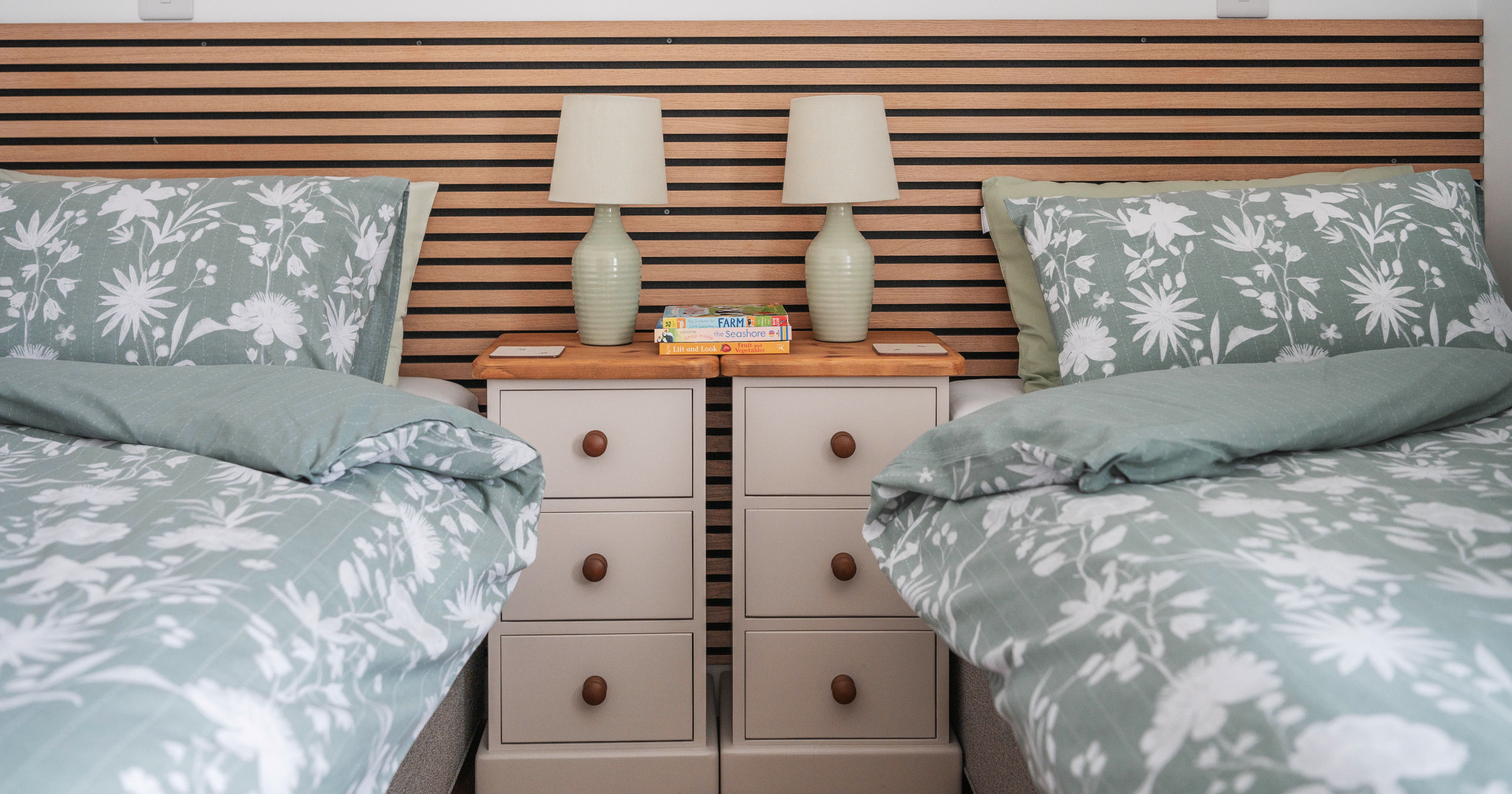 Two twin beds with green floral bedding, a wooden headboard, and a shared nightstand with lamps and books