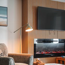 Cozy living room with gray armchair, ottoman, wall-mounted TV, electric fireplace, and wooden slat accent wall