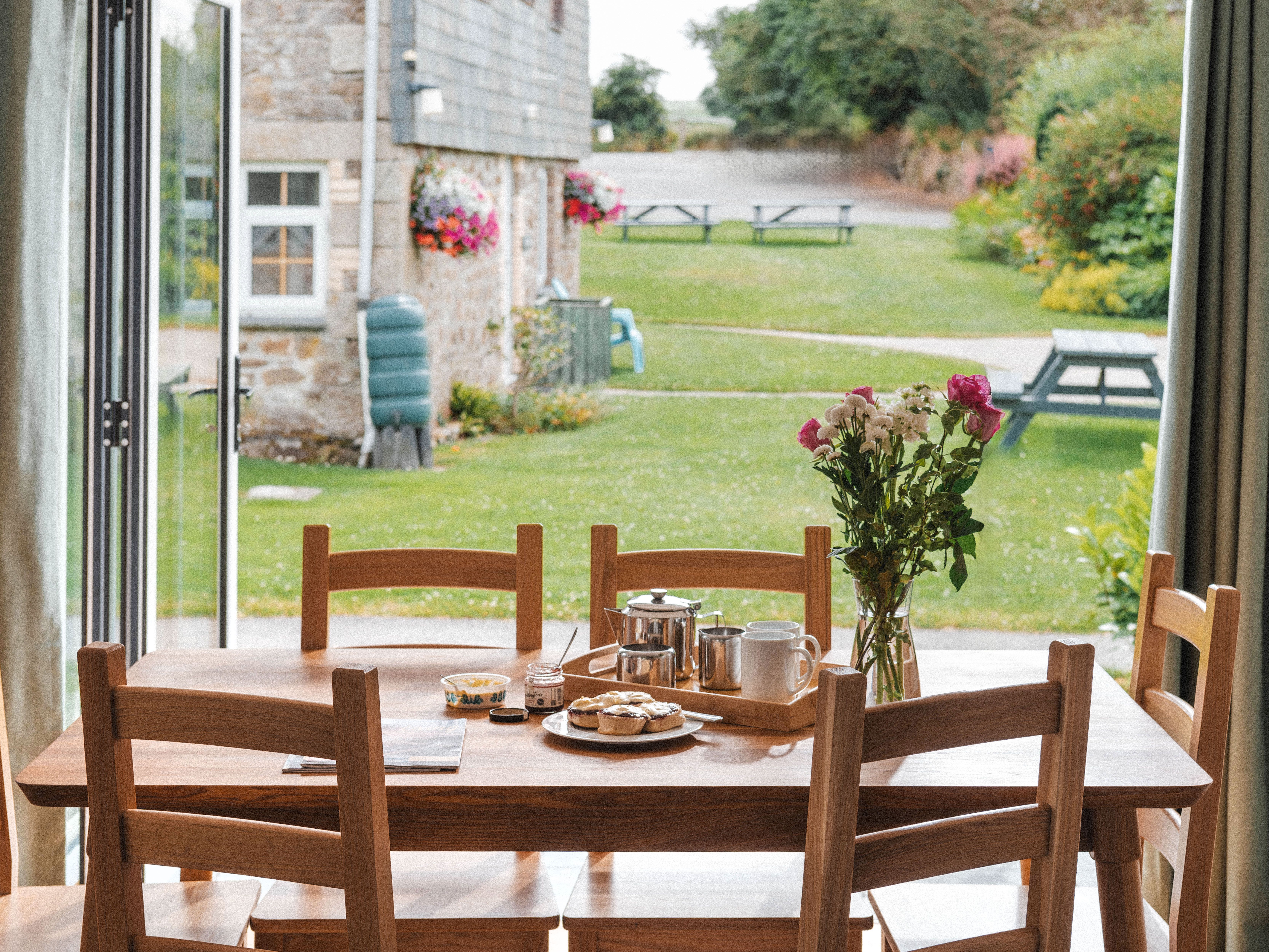 Wooden dining table set for breakfast by open patio doors, overlooking a garden with picnic benches.