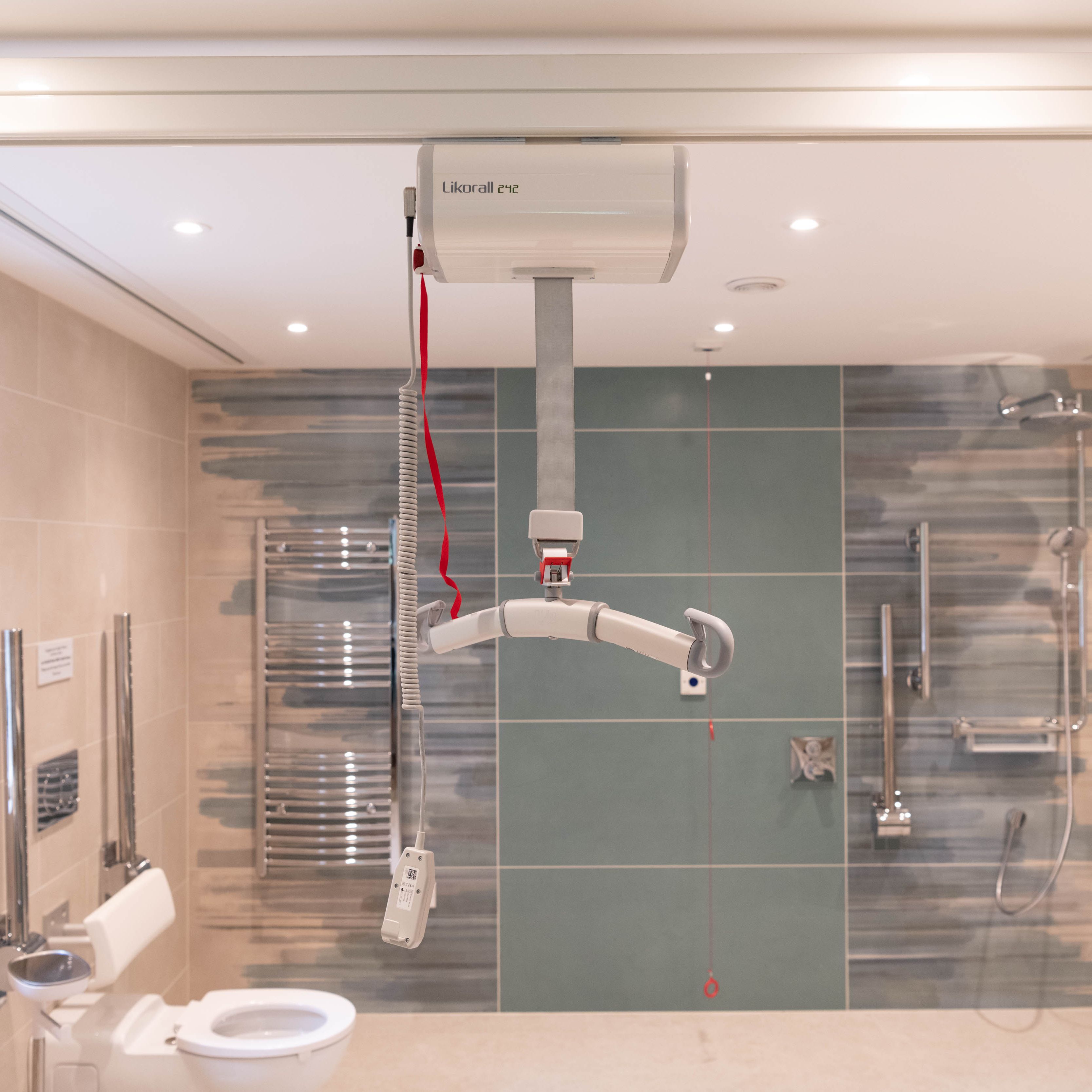Accessible bathroom with a ceiling hoist, foldable shower seat, grab bars, and open shower area.