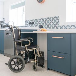 Accessible kitchen with a wheelchair placed by a counter with blue drawers and patterned tiles.