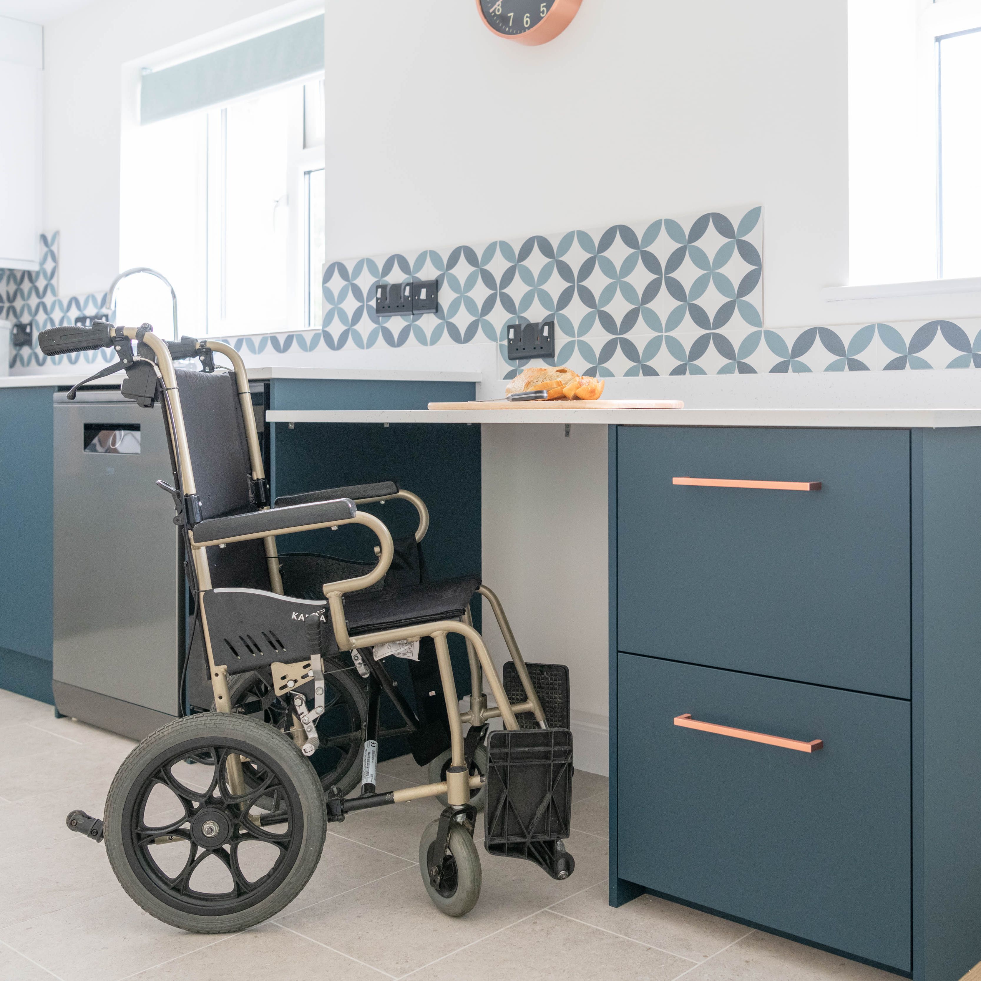 Accessible kitchen with a wheelchair placed by a counter with blue drawers and patterned tiles.