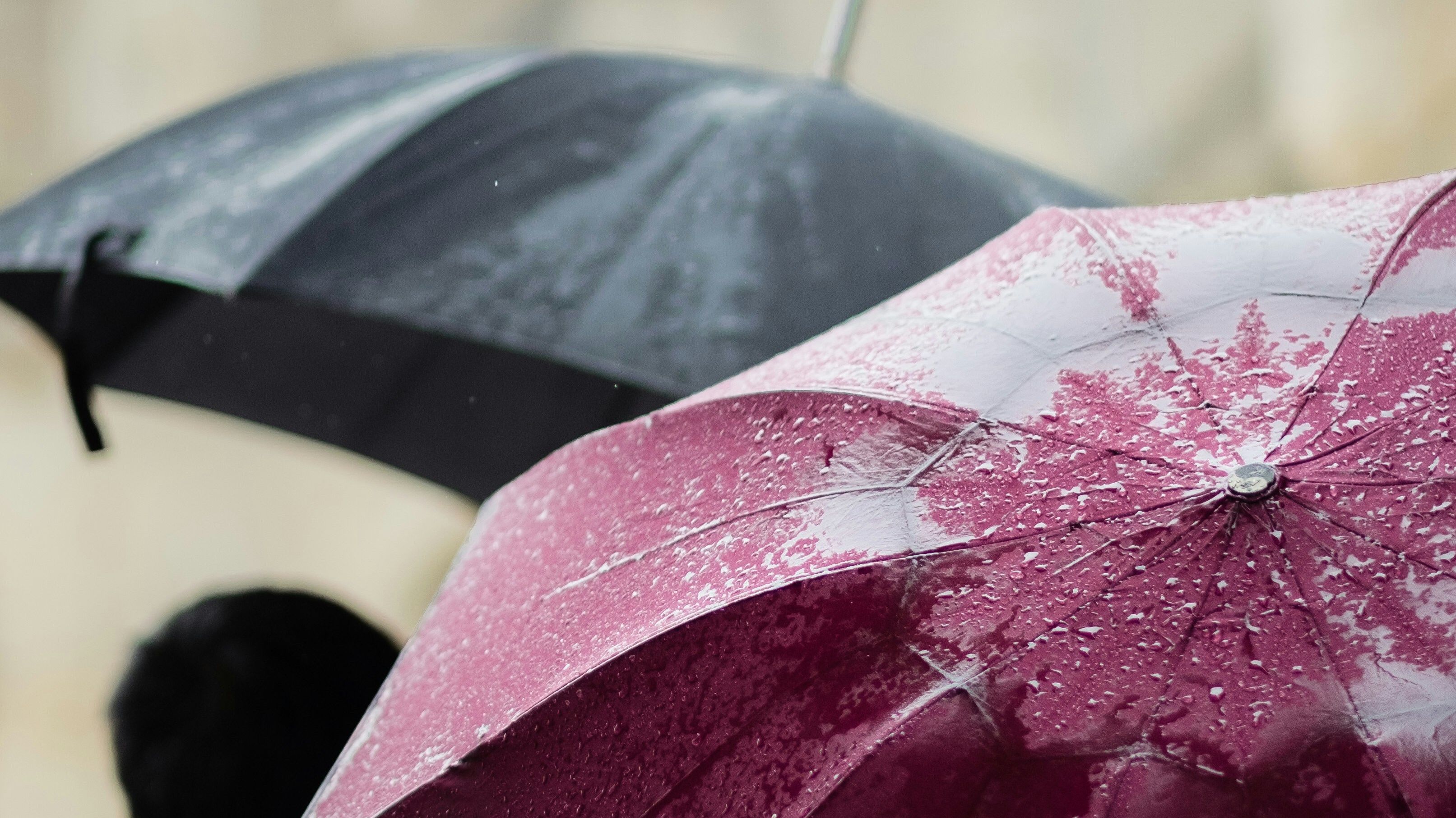 Close-up of a red umbrella covered in raindrops with a person holding a black umbrella in the background