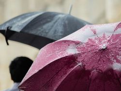 Close-up of a red umbrella covered in raindrops with a person holding a black umbrella in the background