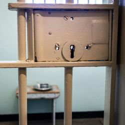 Close-up view of a locked metal jail cell door with part of a cell visible in the background.