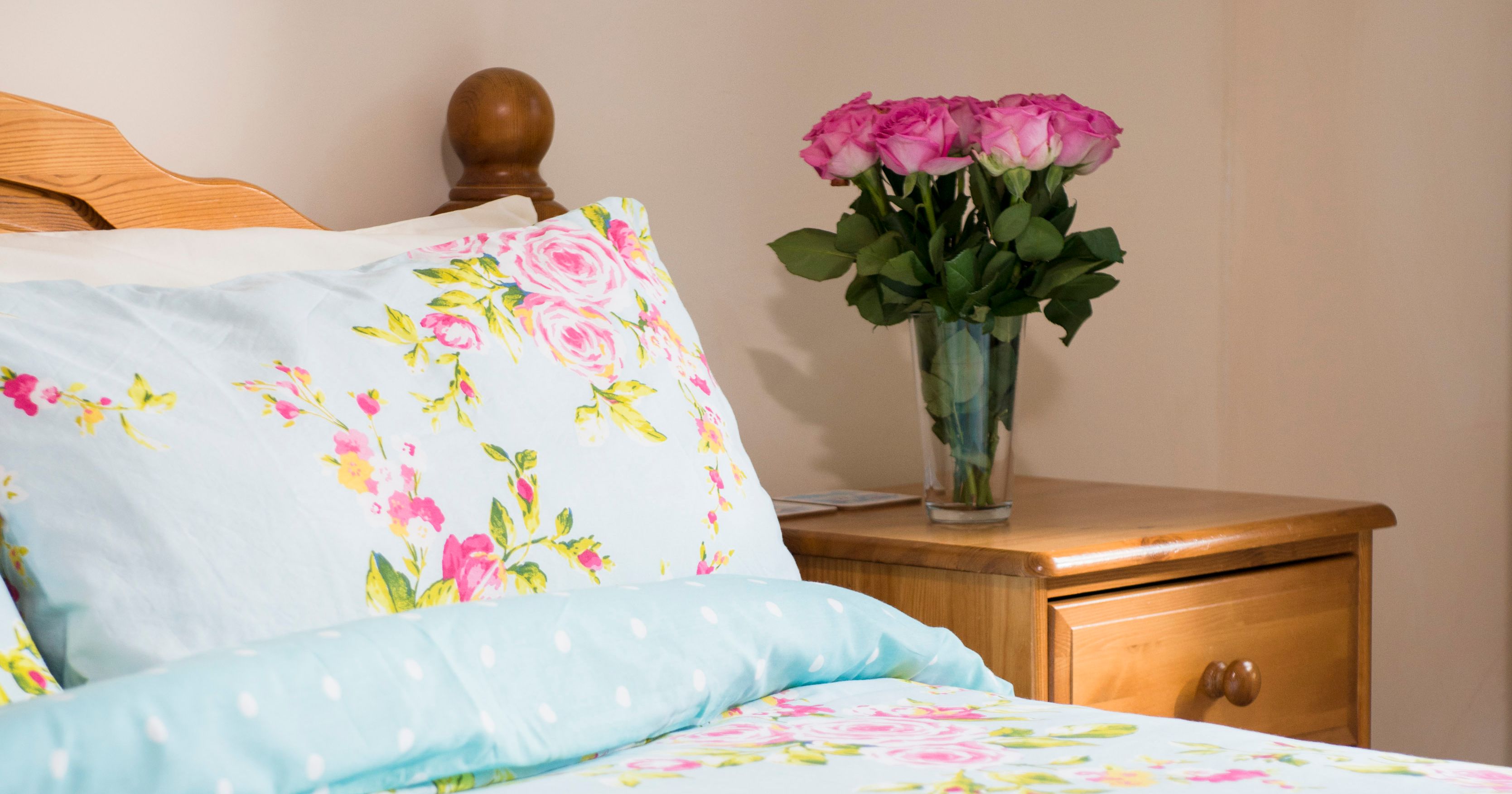 A cozy bedroom with a bed covered in floral bedding, a wooden nightstand, a vase of pink roses, and rolled towels.