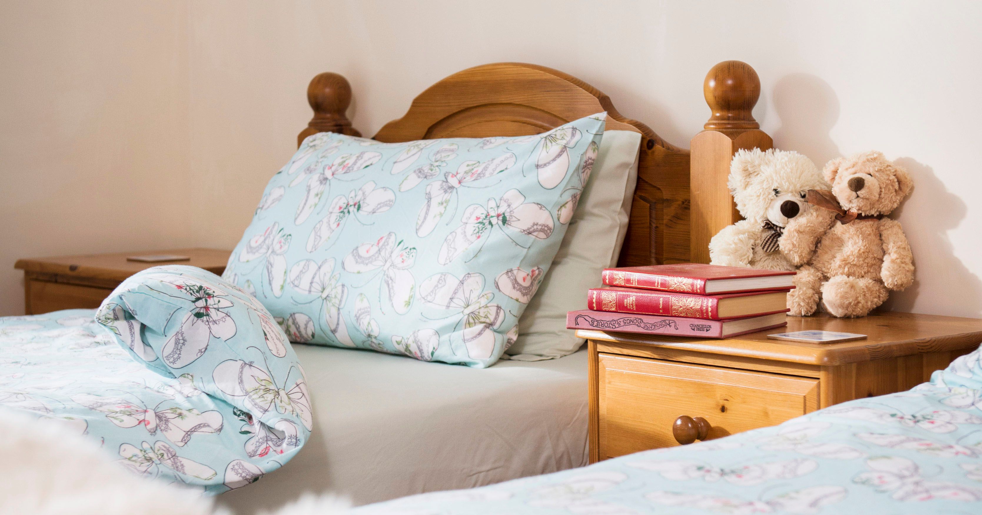 Two single beds with light blue bedding featuring a slippers pattern, a wooden nightstand with teddy bears and books, and a fluffy white object in the foreground.