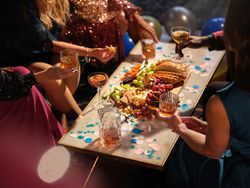 People enjoying drinks and snacks around a festive table with confetti and balloons.