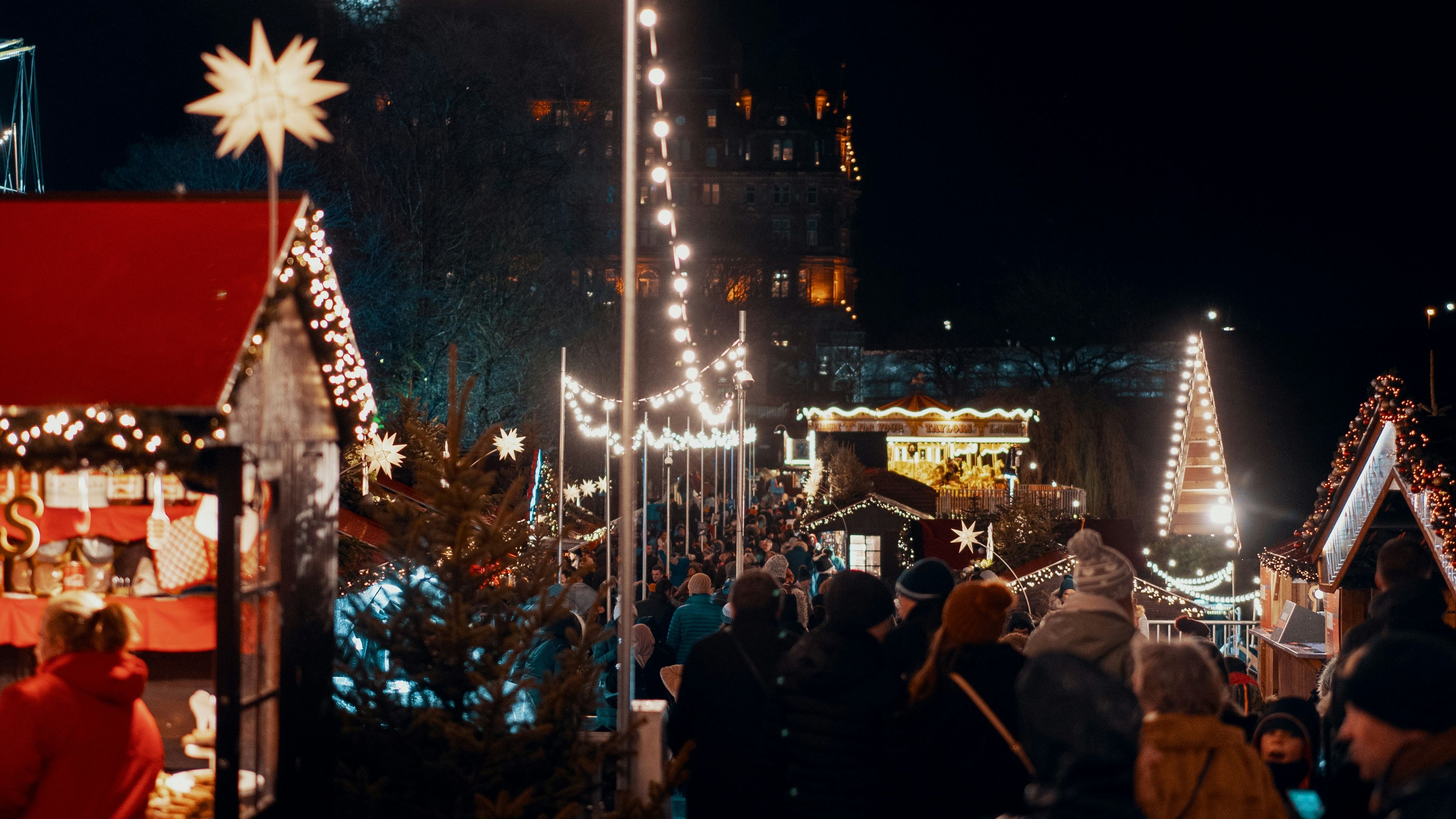 Crowd of people walking through a festive outdoor Christmas market at night with holiday lights and decorations.