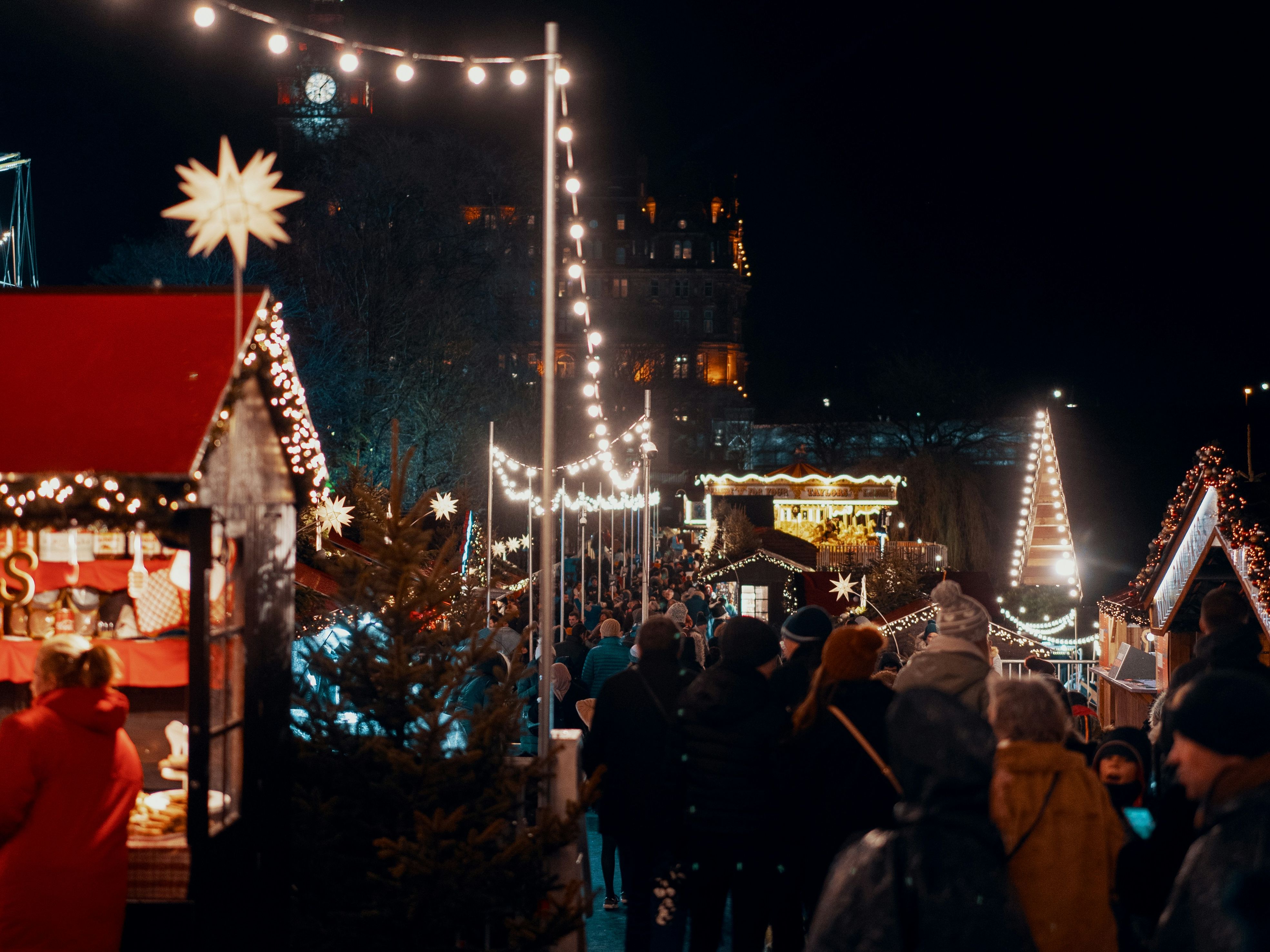 Crowd of people walking through a festive outdoor Christmas market at night with holiday lights and decorations.