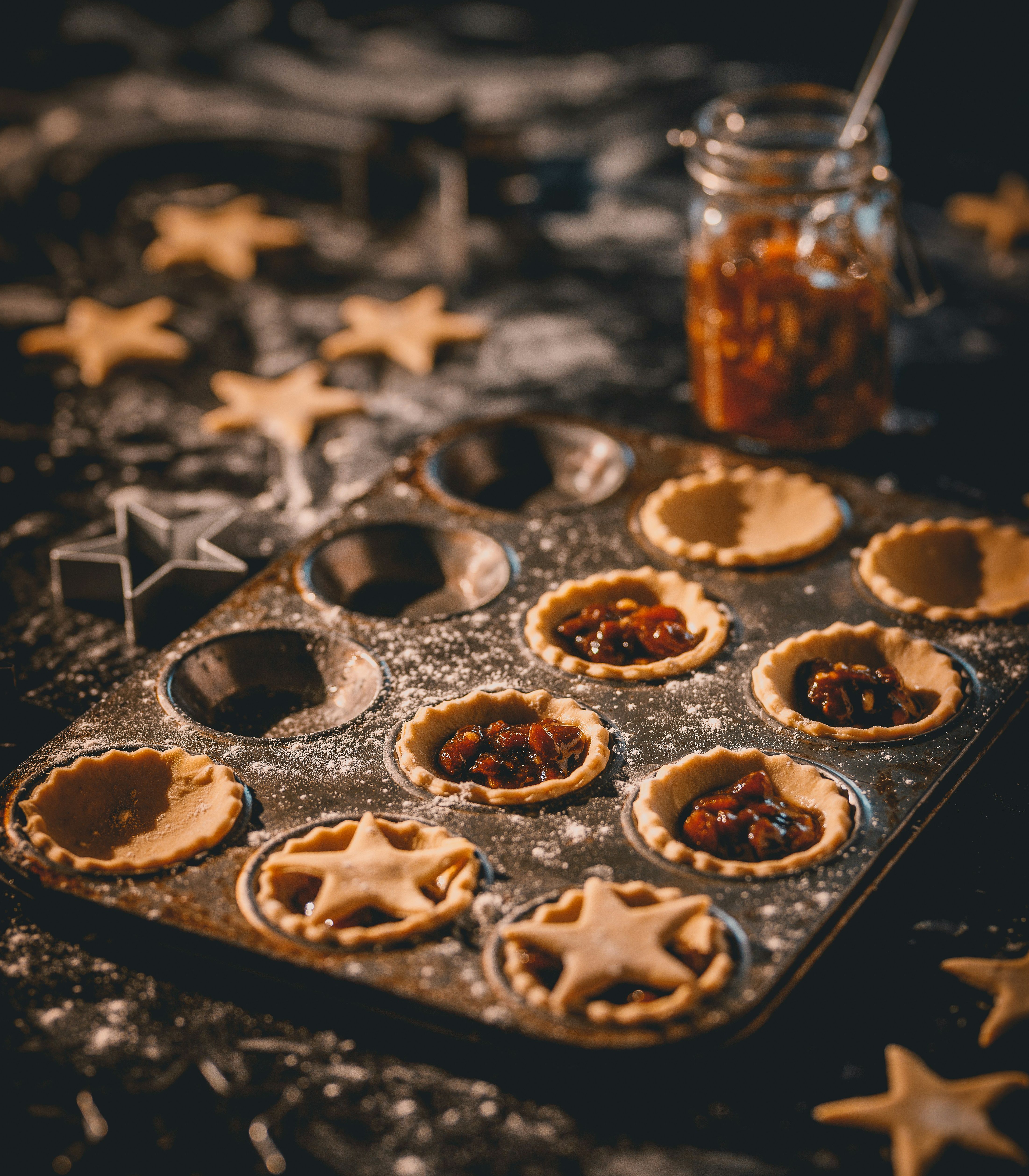 Mince pies being prepared in a baking tray with star-shaped pastry cutouts and a jar of mincemeat in the background.