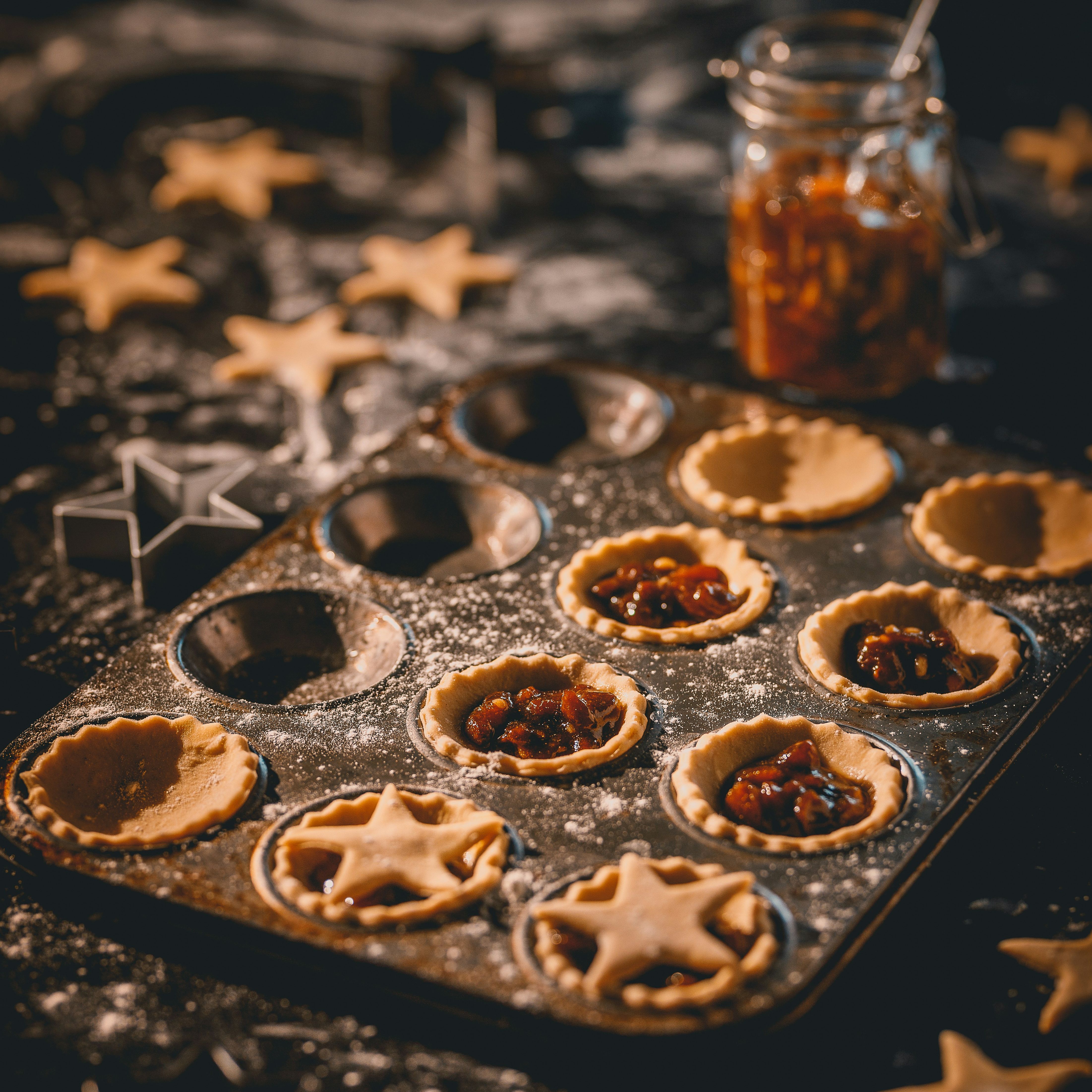 Mince pies being prepared in a baking tray with star-shaped pastry cutouts and a jar of mincemeat in the background.