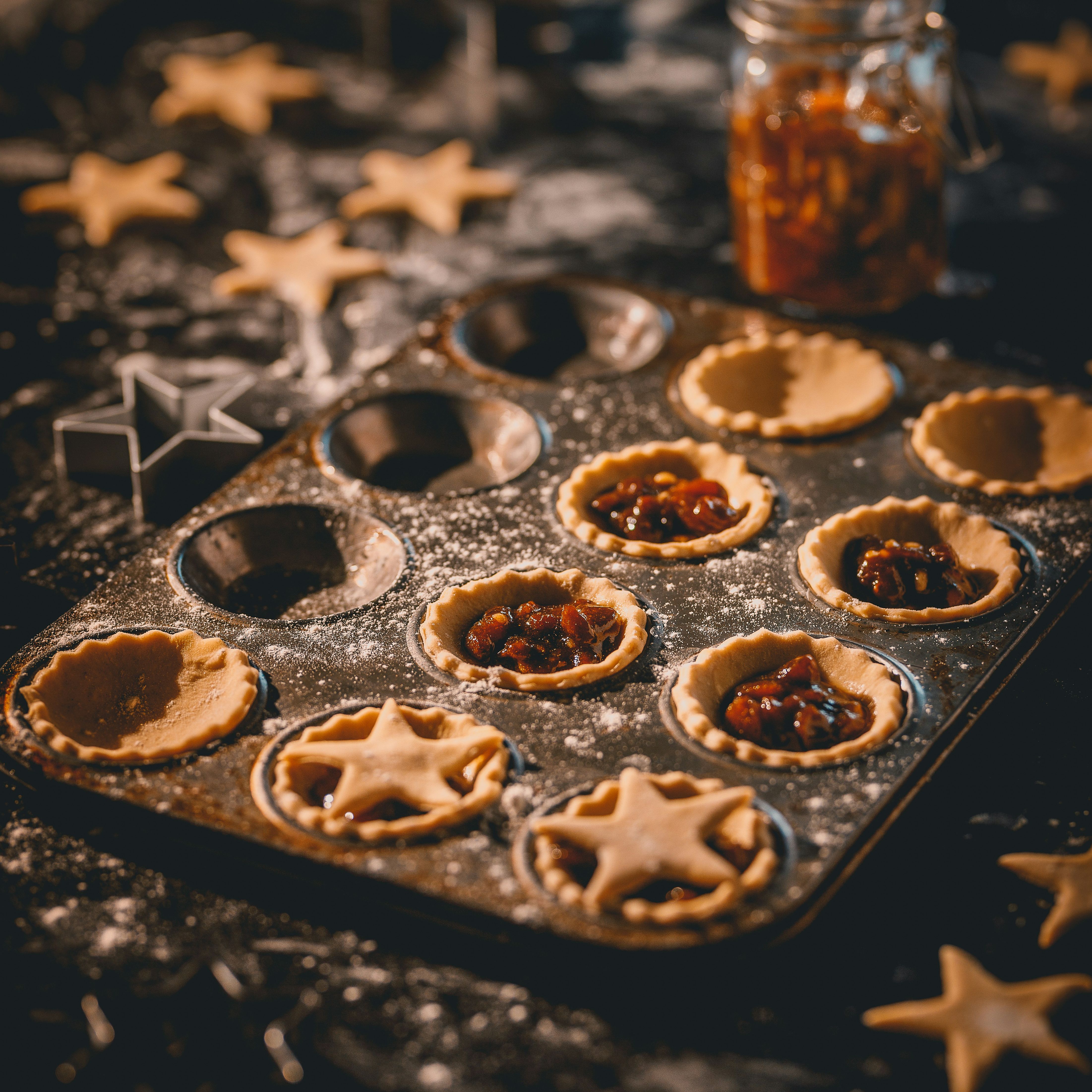 Mince pies being prepared in a baking tray with star-shaped pastry cutouts and a jar of mincemeat in the background.