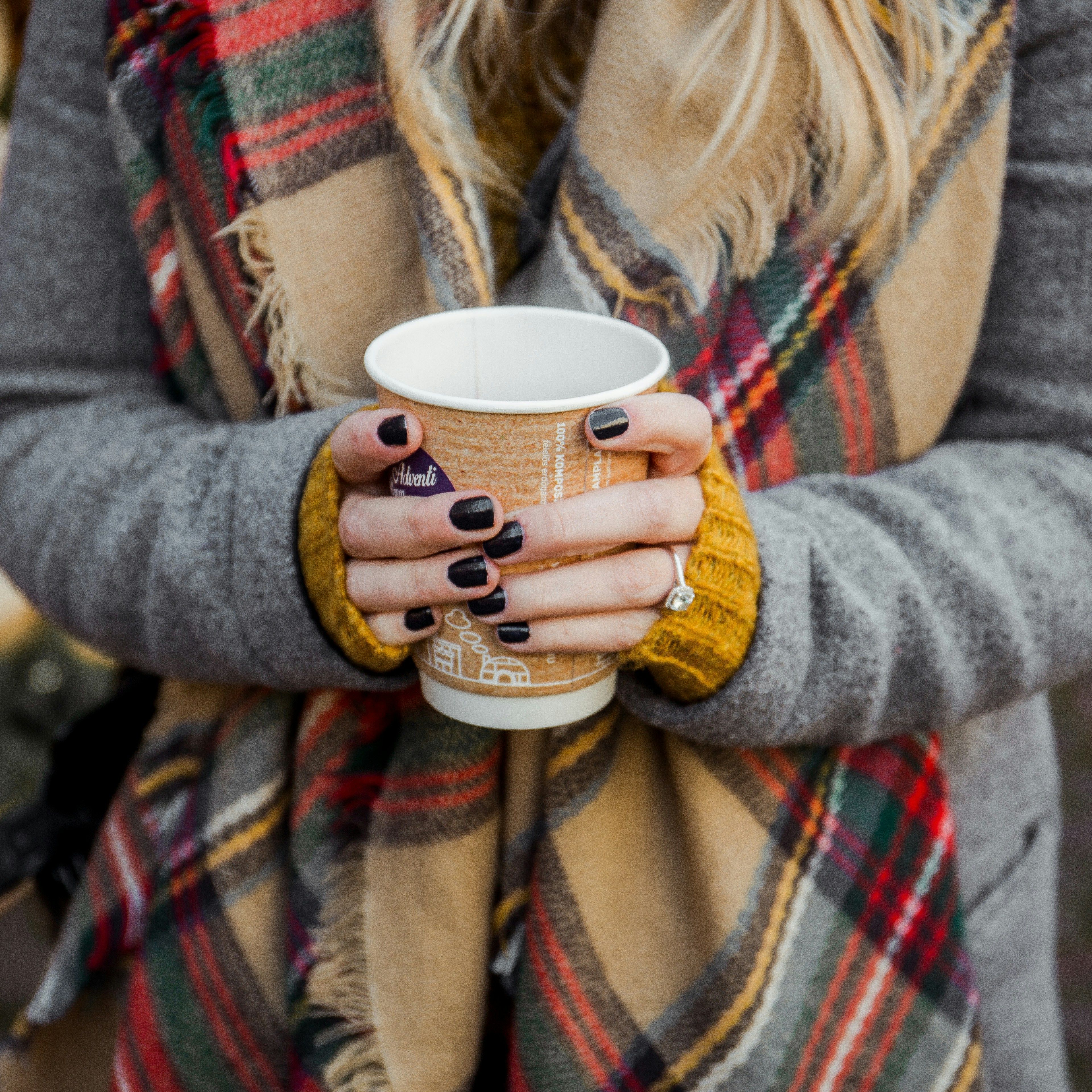 Woman holding a paper coffee cup with both hands while wearing a plaid scarf and gray coat