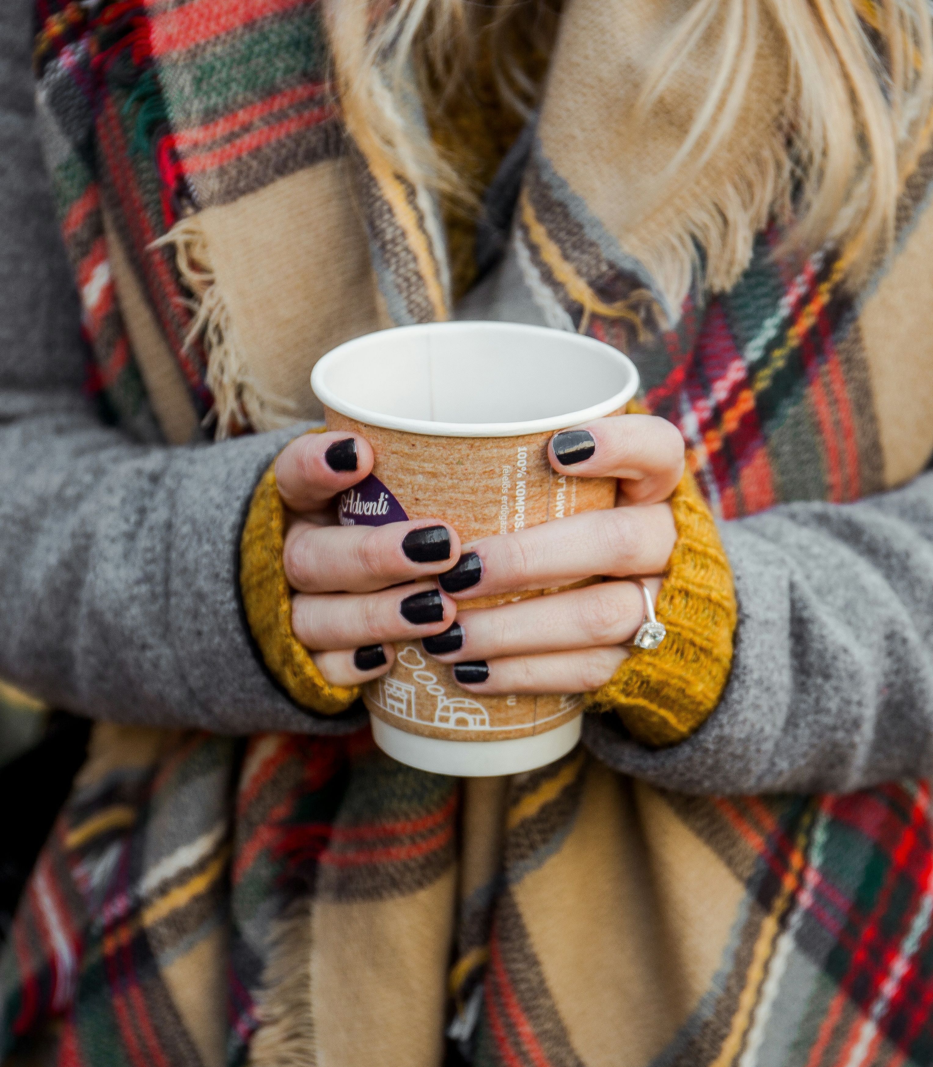 Woman holding a paper coffee cup with both hands while wearing a plaid scarf and gray coat