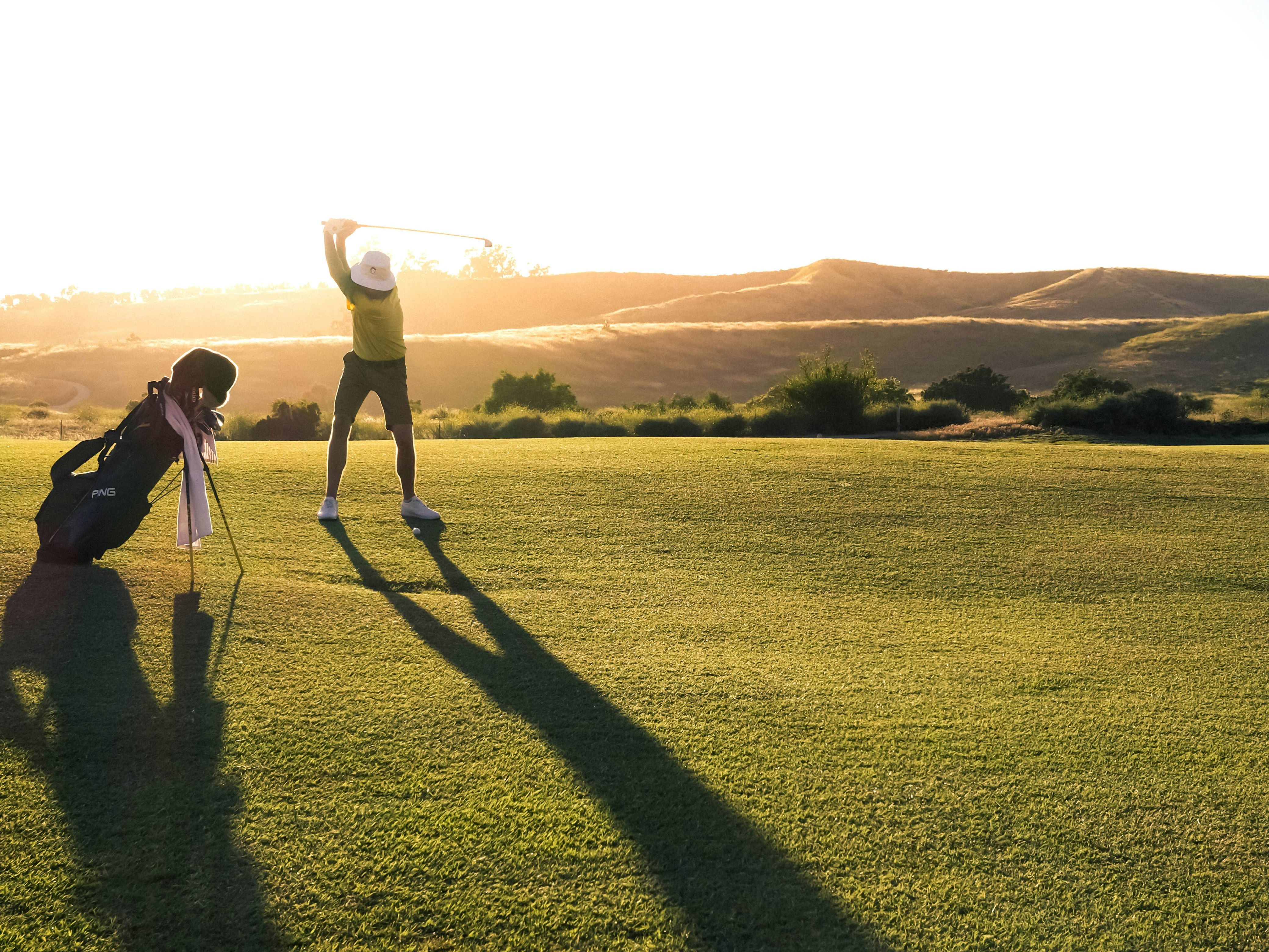 Person preparing to swing a golf club on a golf course at sunset with long shadows cast on the grass