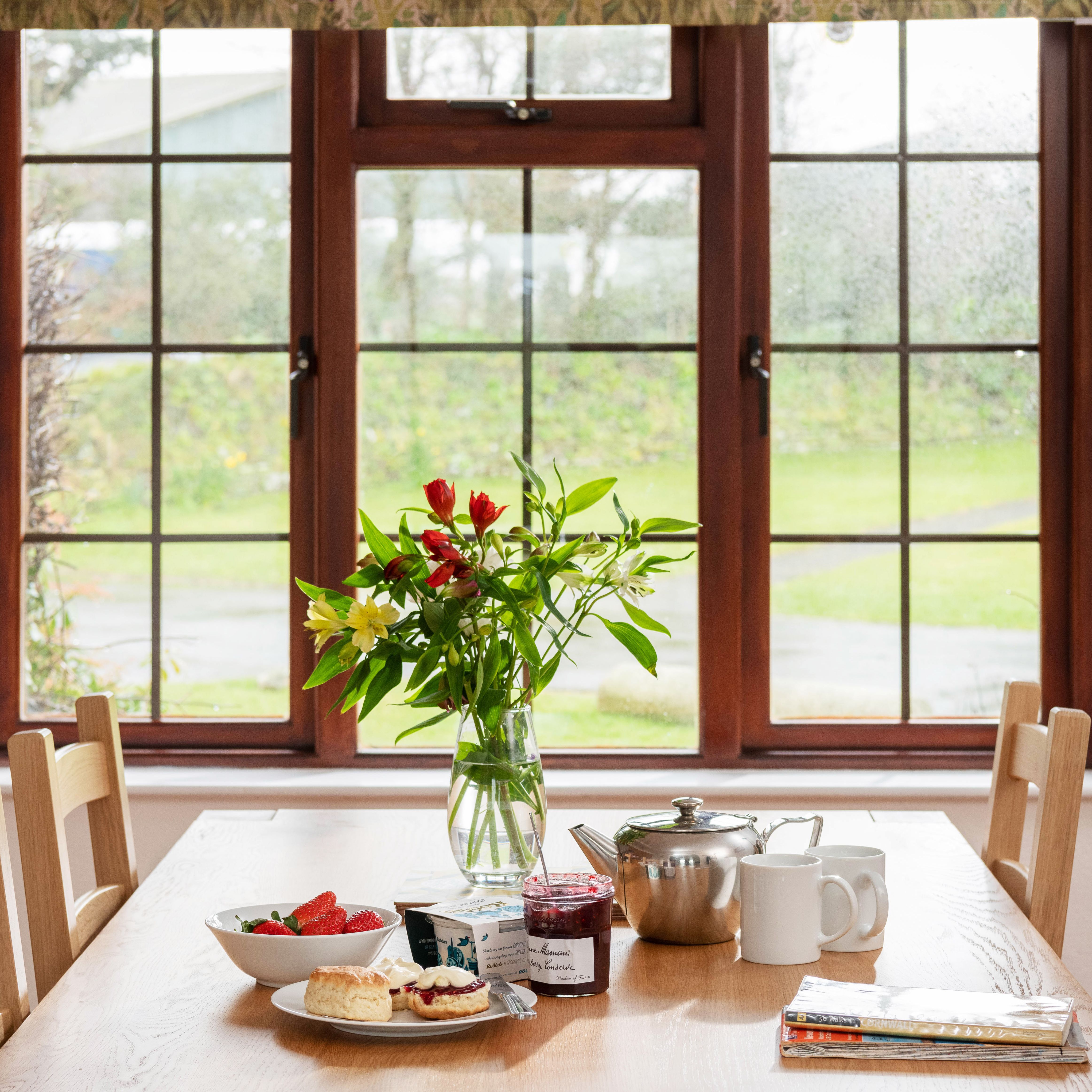 A breakfast table set with tea, jam, scones, a bowl of strawberries, and a vase of flowers in front of a large window.
