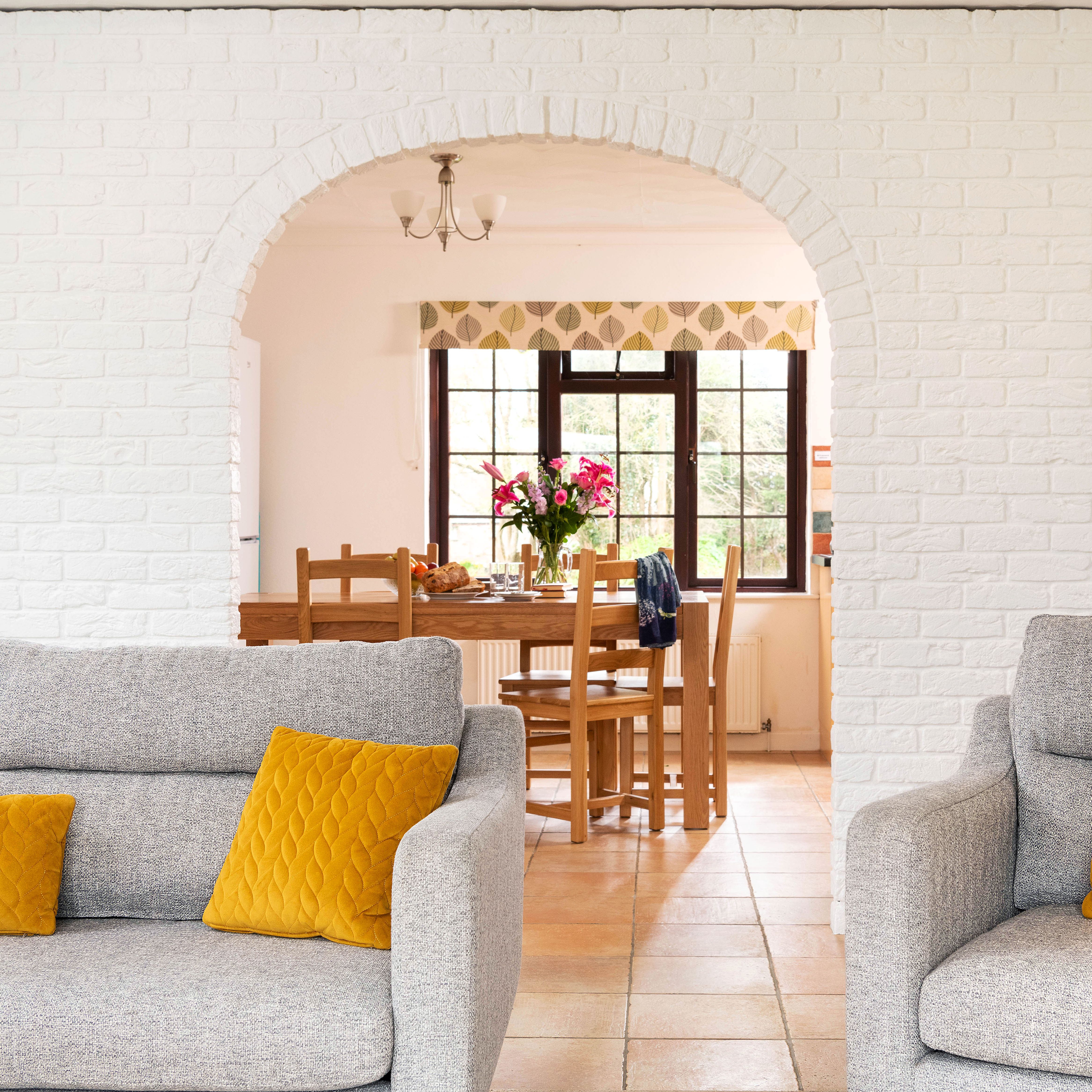 Modern living room with grey sofas and yellow cushions, looking through an archway to a dining table with flowers on it.