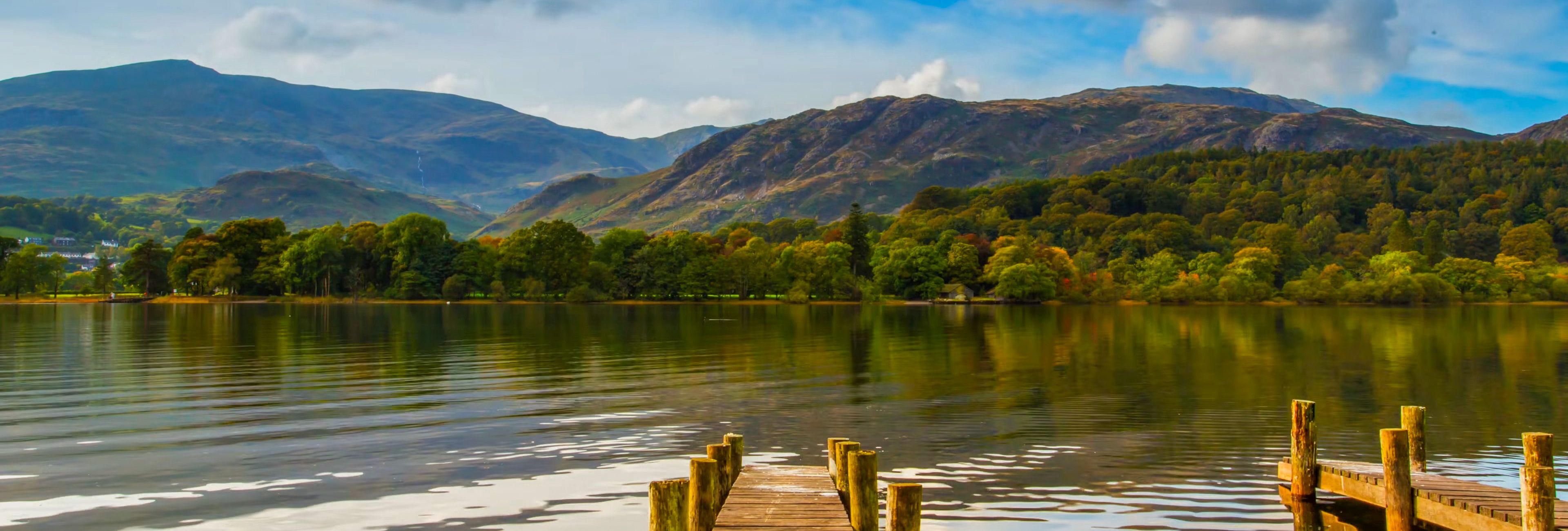 A wooden dock extends into a calm lake, surrounded by lush green trees and rolling hills. The scene is set under a partly cloudy sky with reflections on the water.