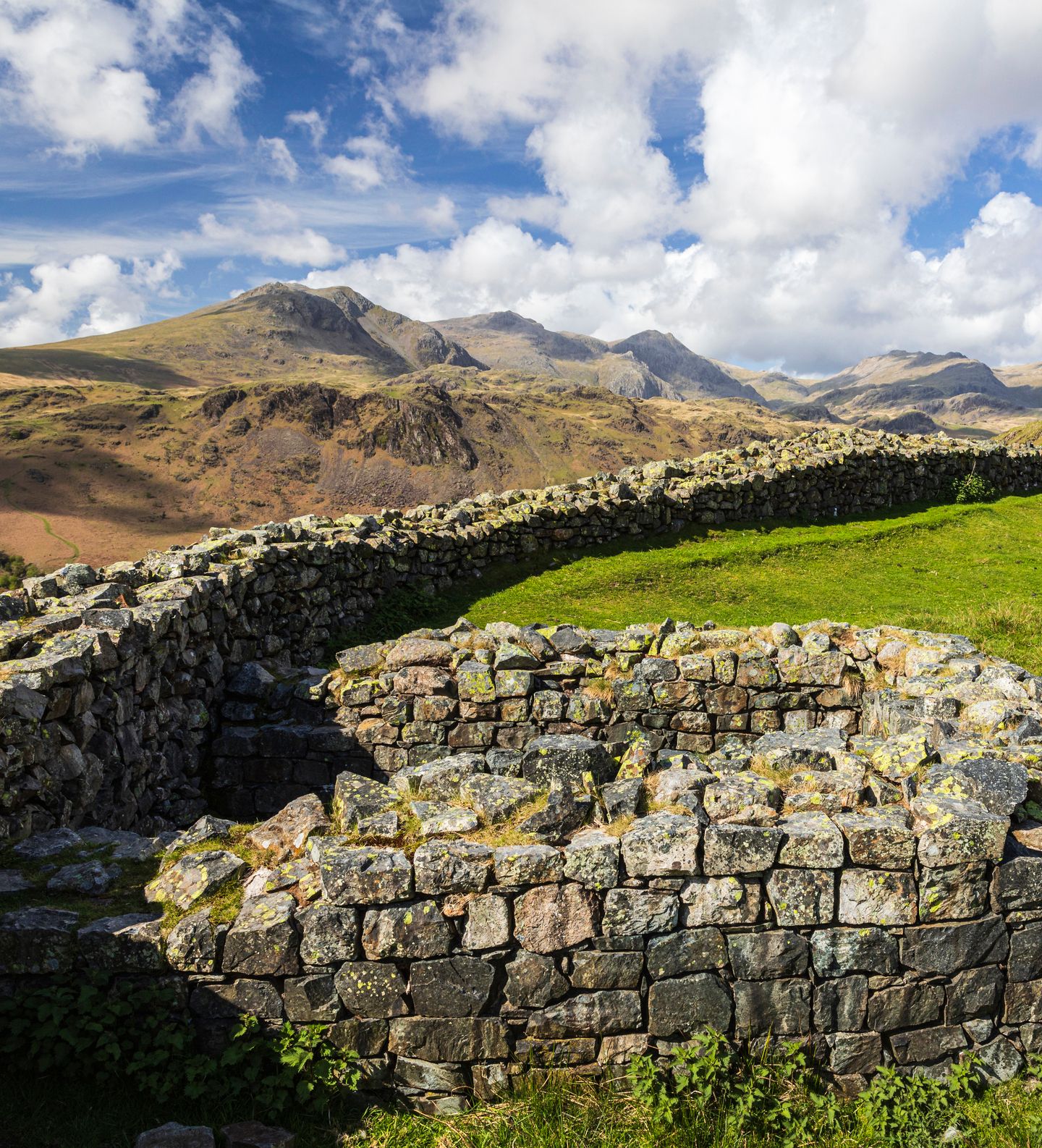 Ancient stone ruins are set against a backdrop of rolling hills and distant mountains under a partly cloudy sky. The grassy landscape contrasts with the weathered stone walls.