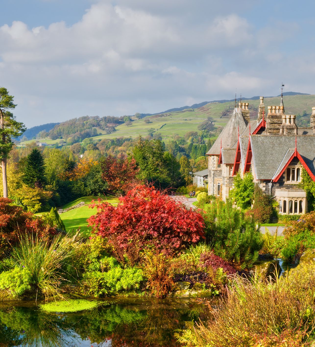 A picturesque estate with a stone manor house, surrounded by vibrant autumn foliage and a reflective pond in the foreground. Rolling green hills stretch into the background.