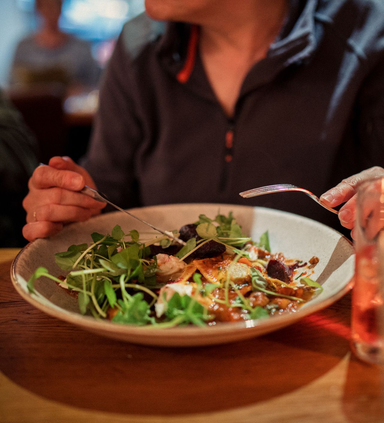 A person is dining in a cozy restaurant, eating a salad with greens and roasted vegetables from a large bowl. Glasses with beverages are placed on the wooden table nearby.