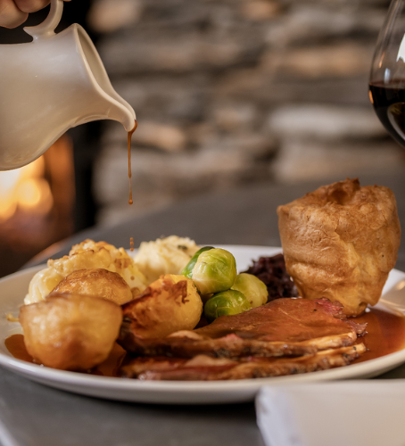 A plate of roast dinner with sliced meat, crispy potatoes, mashed potatoes, Brussels sprouts, and a Yorkshire pudding, as rich gravy is poured from a jug.
