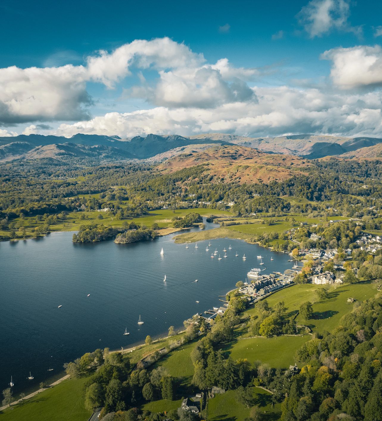 Aerial view of a lake with sailboats, surrounded by green hills, a small town, and distant mountains under a blue sky with scattered clouds.
