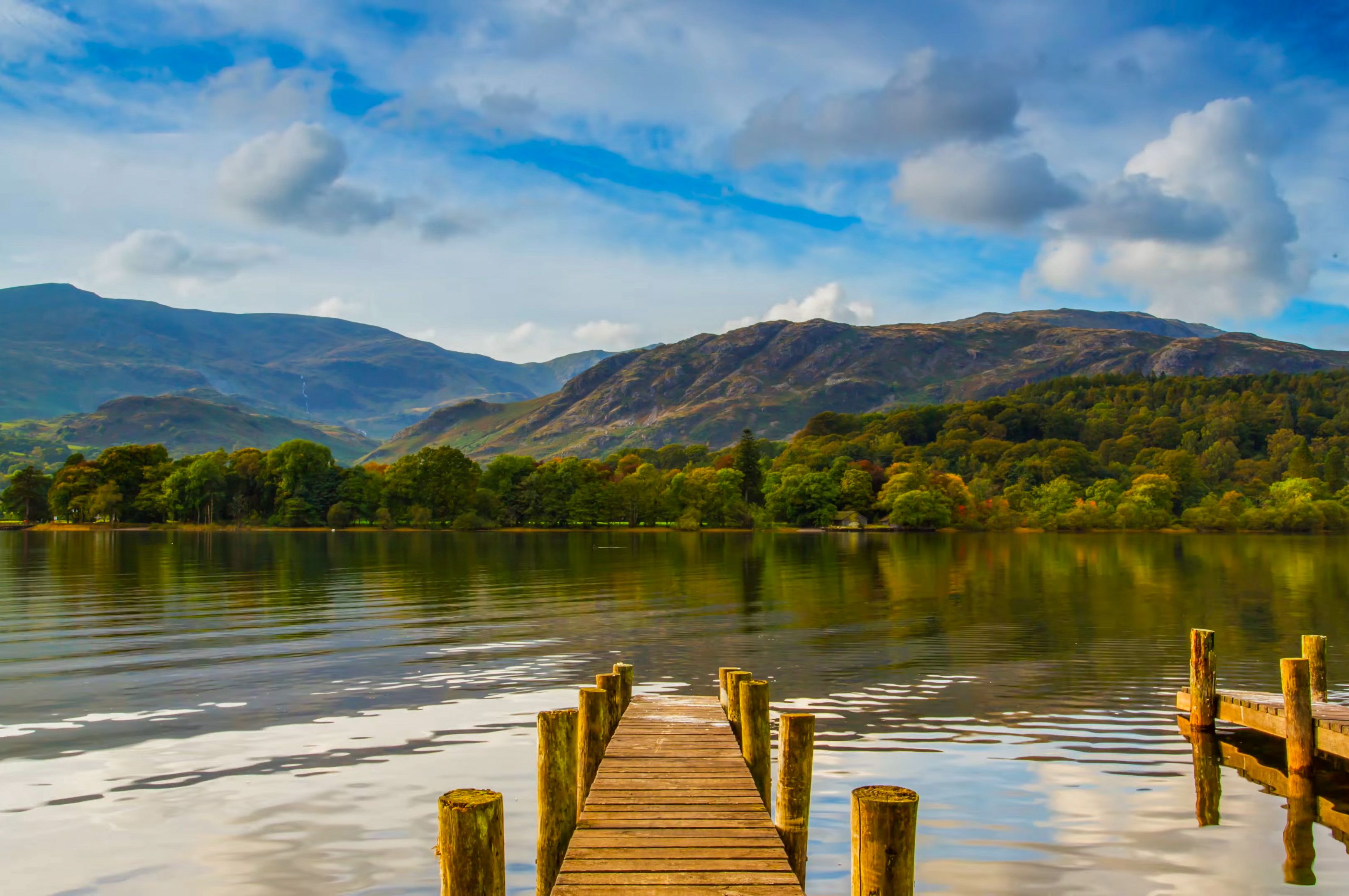 A wooden pier extends into a calm lake, reflecting the surrounding green hills and distant mountains under a partly cloudy blue sky.