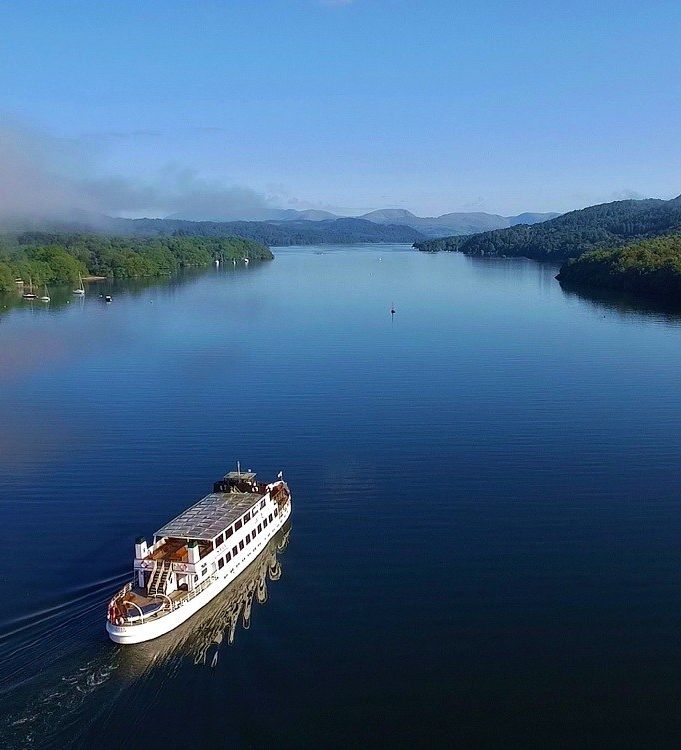 A white passenger boat cruises across a calm, wide lake surrounded by forested hills and distant mountains under a clear blue sky.