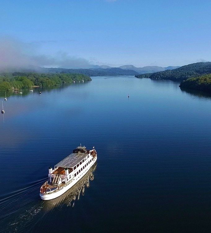 A white passenger boat cruises across a calm, wide lake surrounded by forested hills and distant mountains under a clear blue sky.