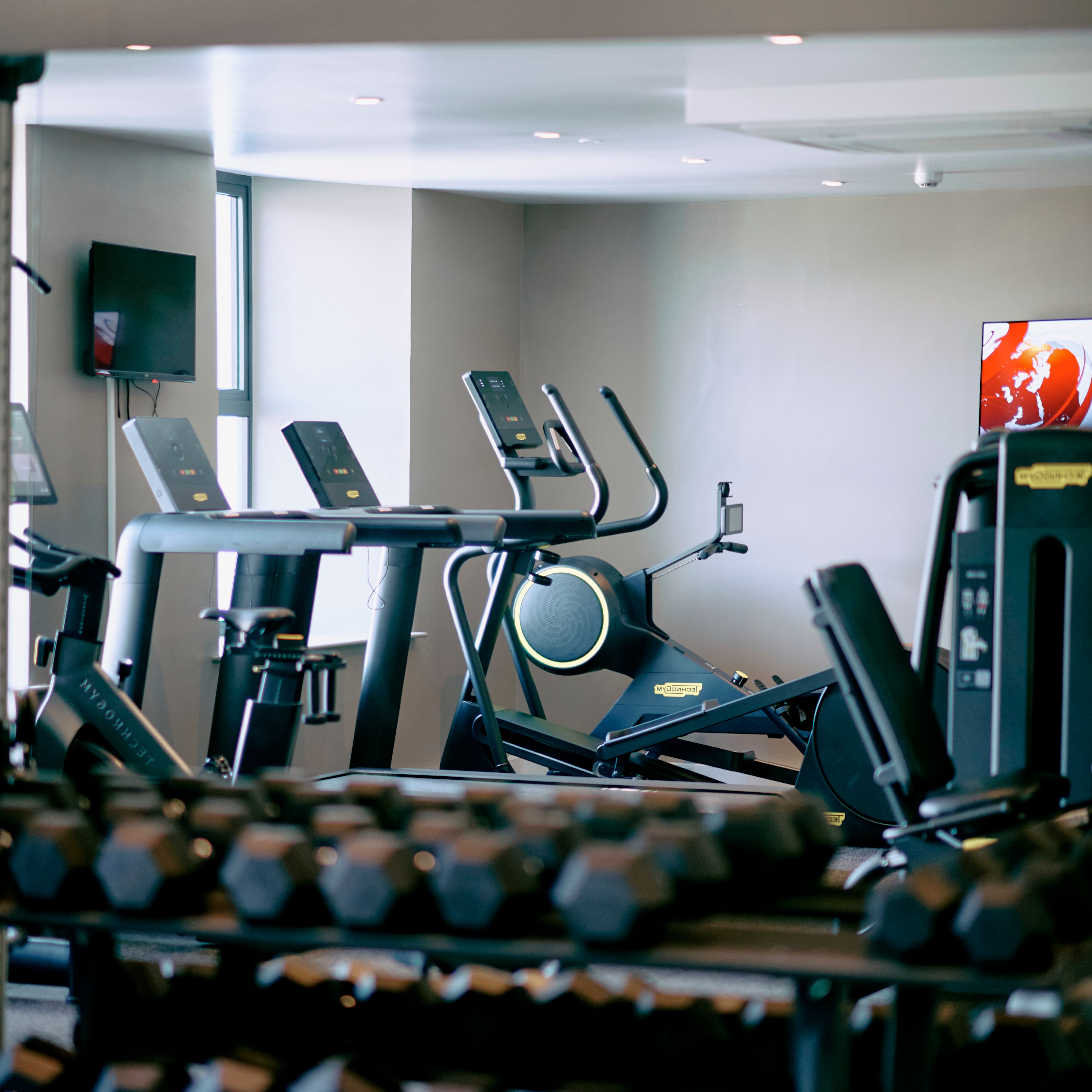A modern gym with rows of dumbbells in the foreground and various cardio machines, including treadmills and an elliptical, visible in the background.