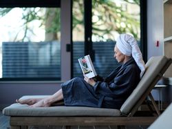 A woman in a bathrobe and towel turban relaxes on a lounge chair indoors, reading a book with a flower vase on the cover, beside large windows.