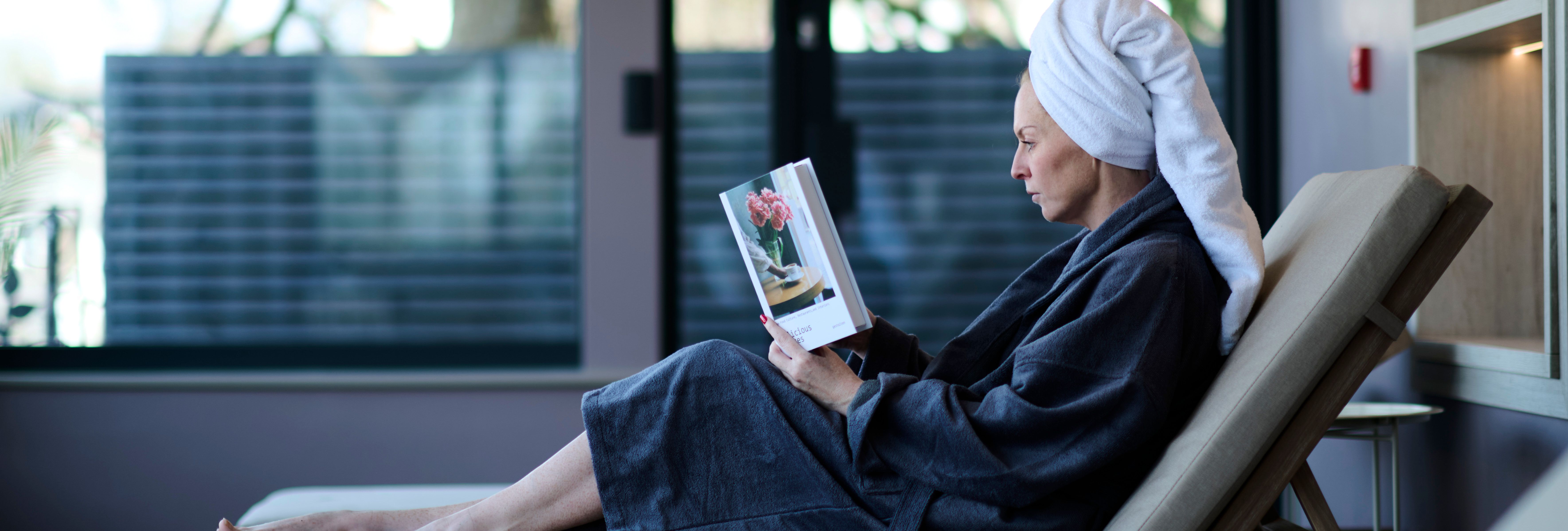 A woman in a bathrobe and towel turban relaxes on a lounge chair indoors, reading a book with a flower vase on the cover, beside large windows.