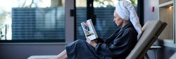 A woman in a bathrobe and towel turban relaxes on a lounge chair indoors, reading a book with a flower vase on the cover, beside large windows.