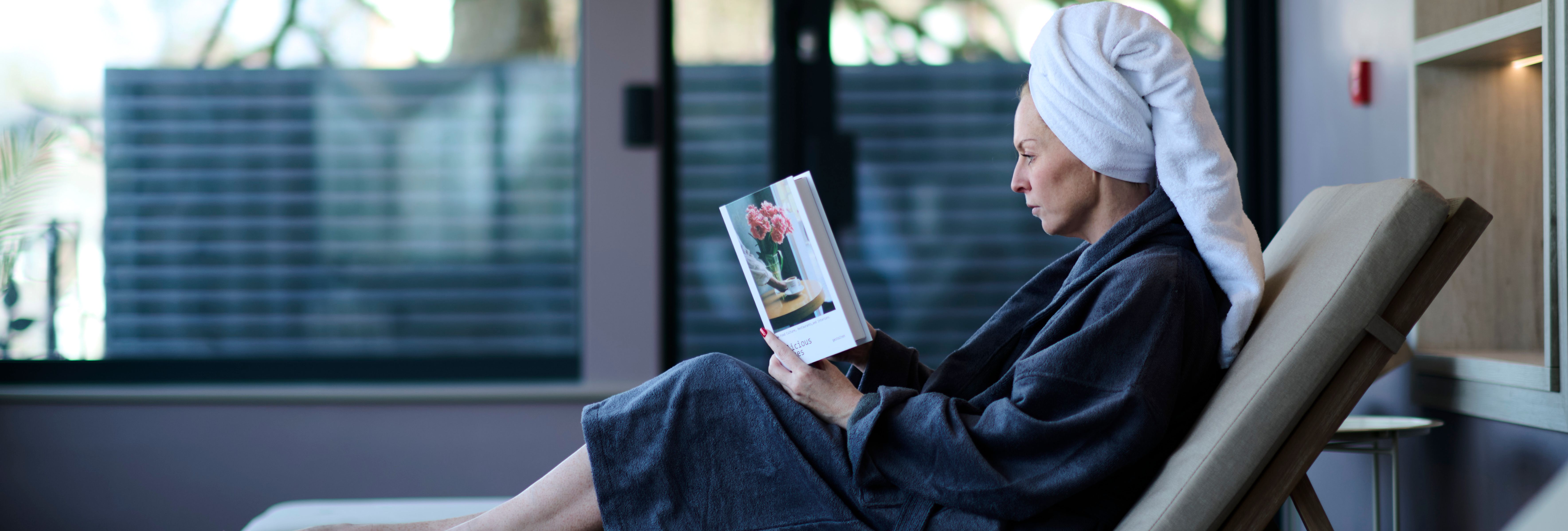 A woman in a bathrobe and towel turban relaxes on a lounge chair indoors, reading a book with a flower vase on the cover, beside large windows.